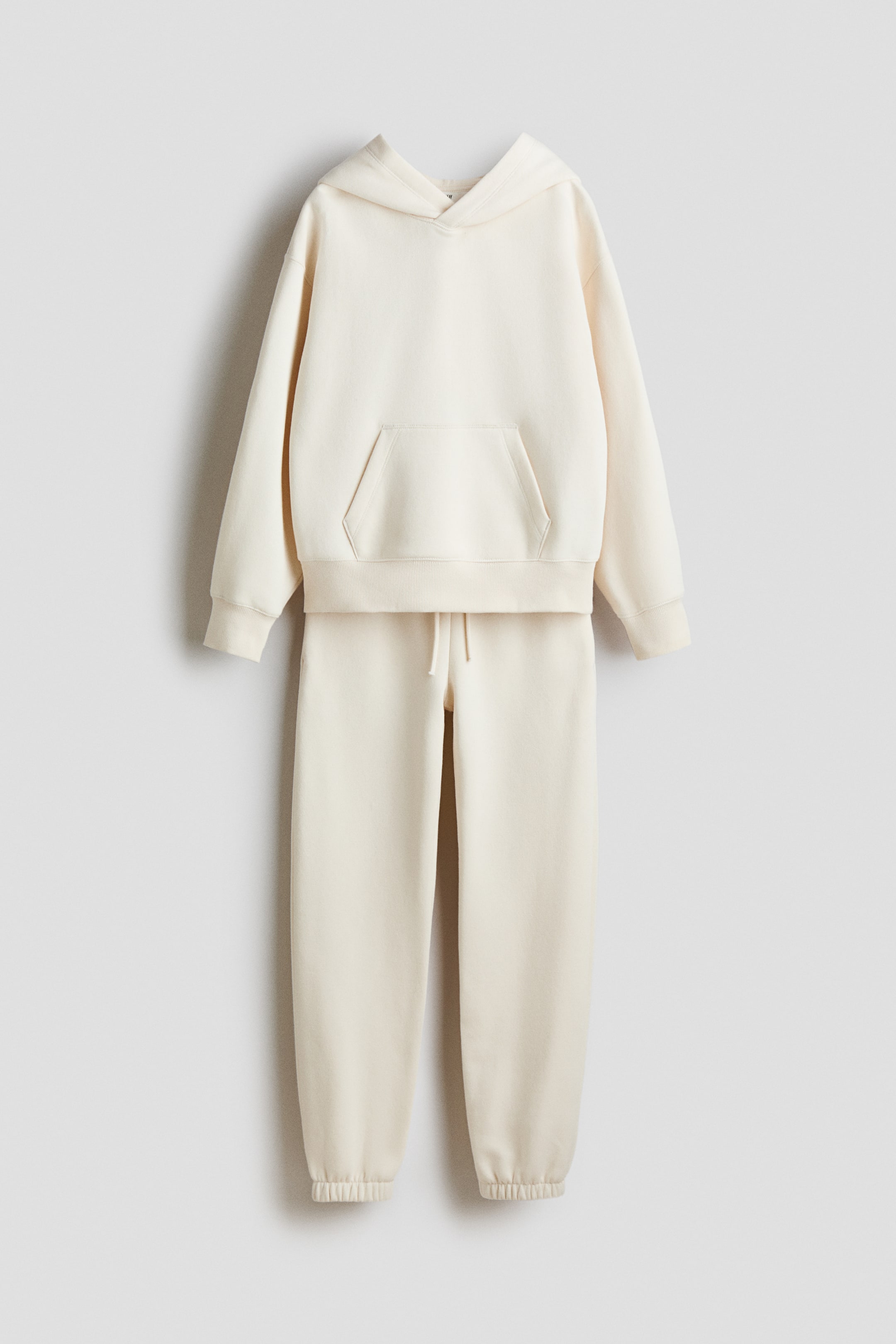 View larger image: A cream-colored tracksuit, viewed from the front, is featuring a matching hoodie and sweatpants. The hoodie is presenting a hood, a front kangaroo pocket, ribbed cuffs, and a ribbed hem, while the sweatpants are displaying an elastic waistband with a visible drawstring and ribbed ankle cuffs.