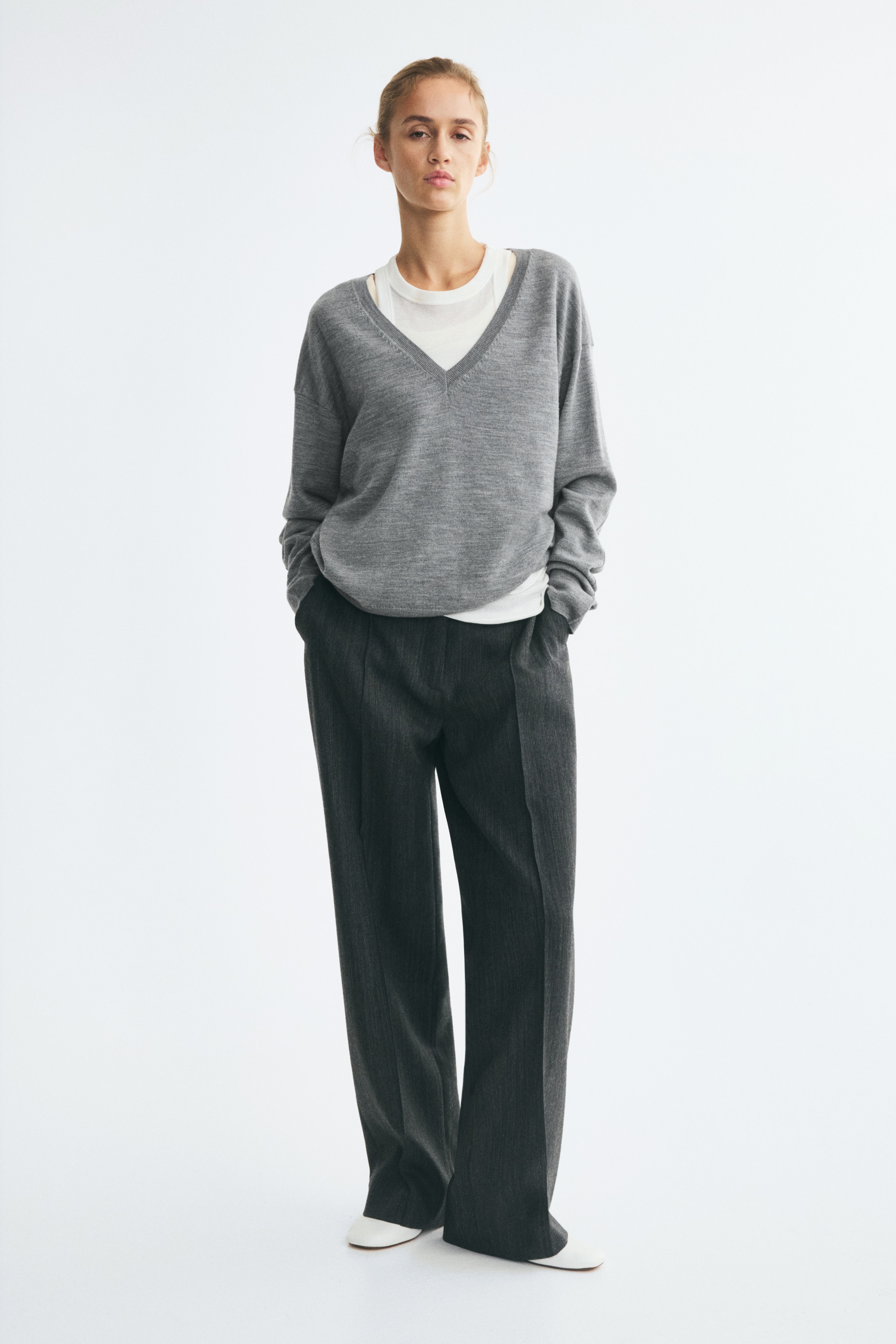 A grey knit sweater is shown from the front, featuring a deep V-neckline with ribbed trim and dropped shoulders. The sweater has a relaxed fit with long sleeves ending in ribbed cuffs and a matching ribbed hem.
