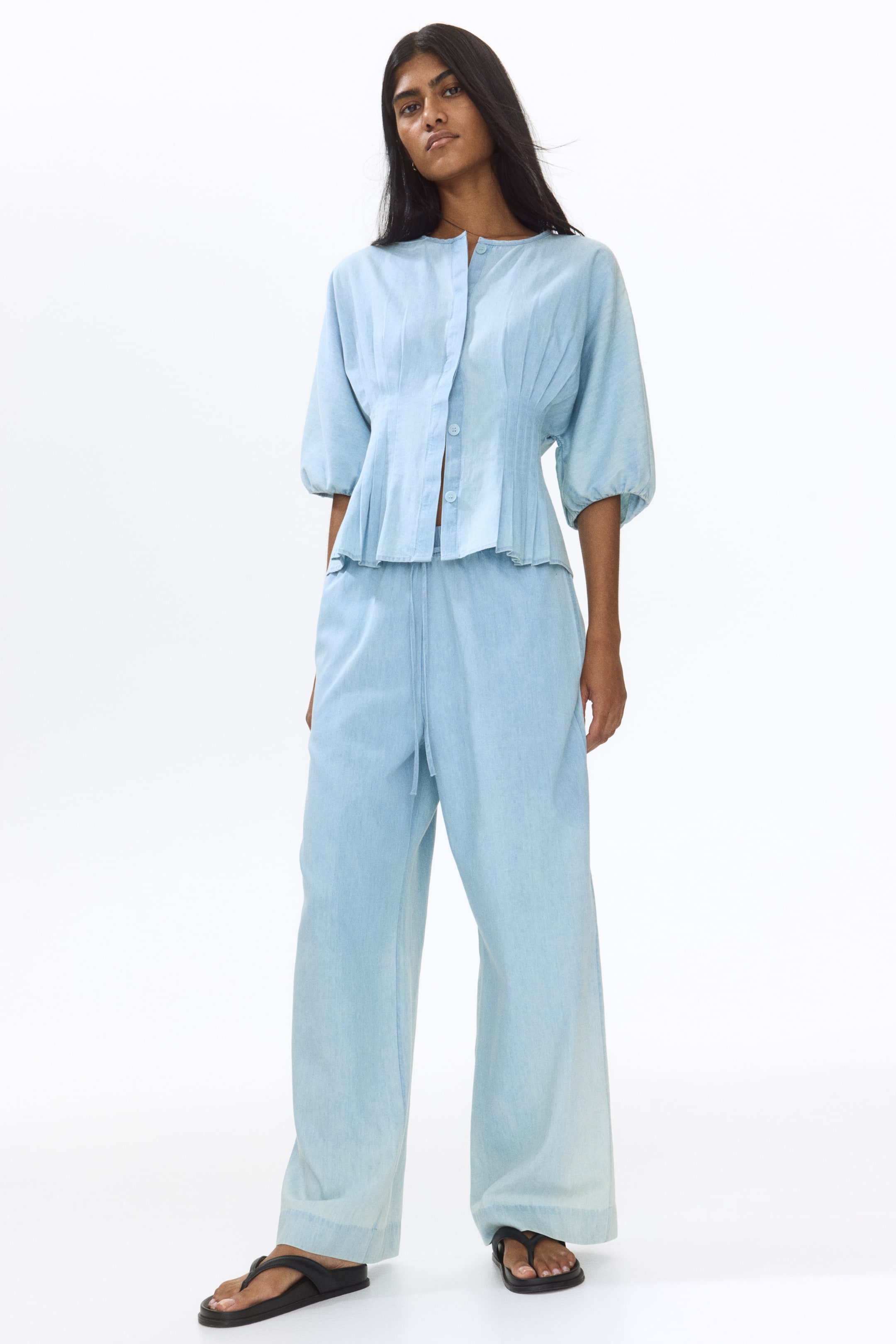 View larger image: A light blue button-front peplum top, featuring a round neck, pin tucks, and voluminous half-sleeves, is showcased from a front angle. It is styled with matching wide-leg drawstring pants that gather at the ankles.