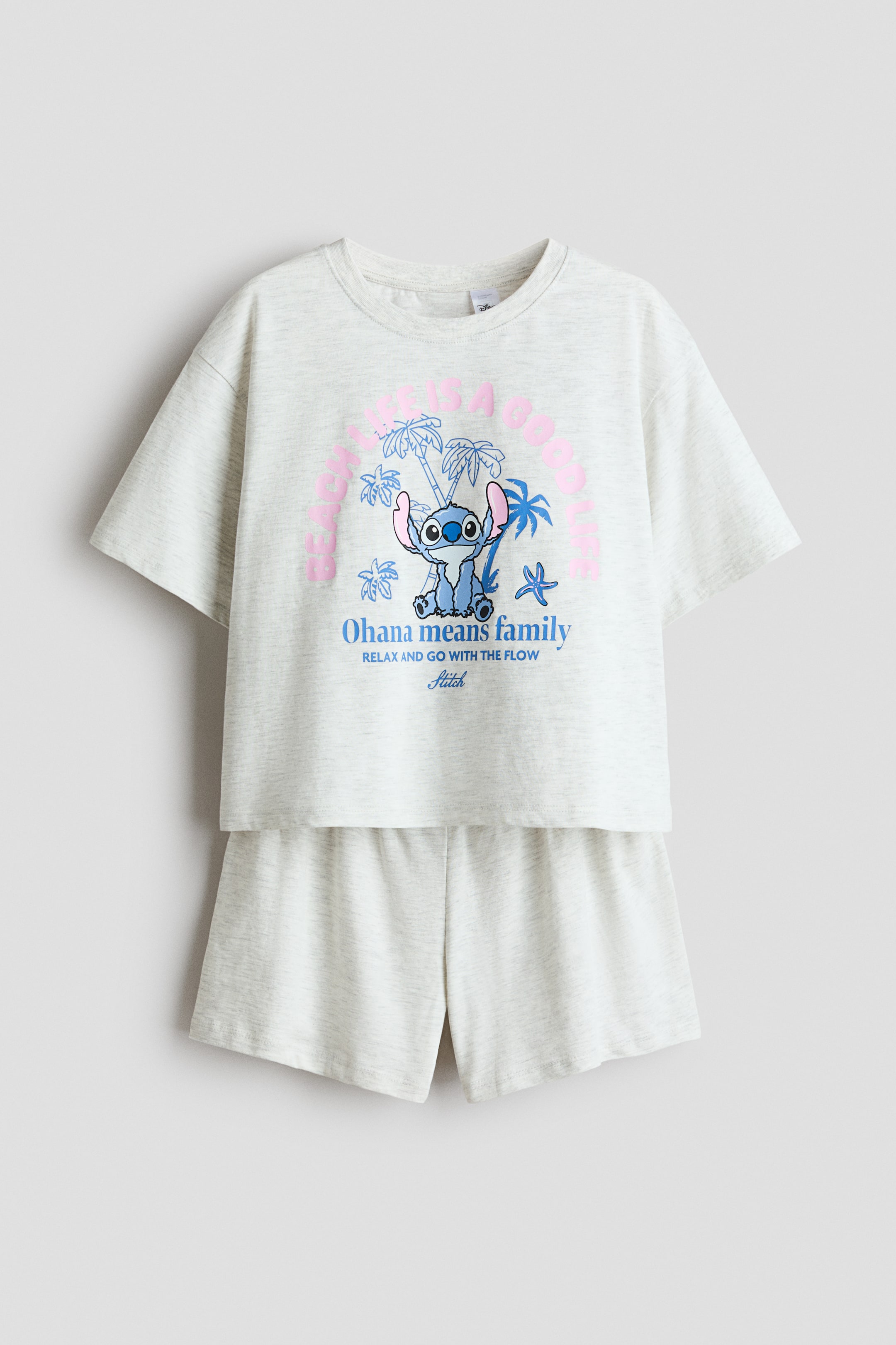 Agrandir l'image: Un ensemble de pyjamas en jersey de coton gris clair chiné présente un T-shirt et un short assorti, vus de face. Le T-shirt arbore une bordure côtelée autour de l'encolure, des épaules tombantes, et un imprimé graphique coloré de Stitch assis parmi des palmiers et des étoiles de mer avec les textes "BEACH LIFE IS A GOOD LIFE" et "OHANA MEANS FAMILY". Le short affiche une taille élastiquée.