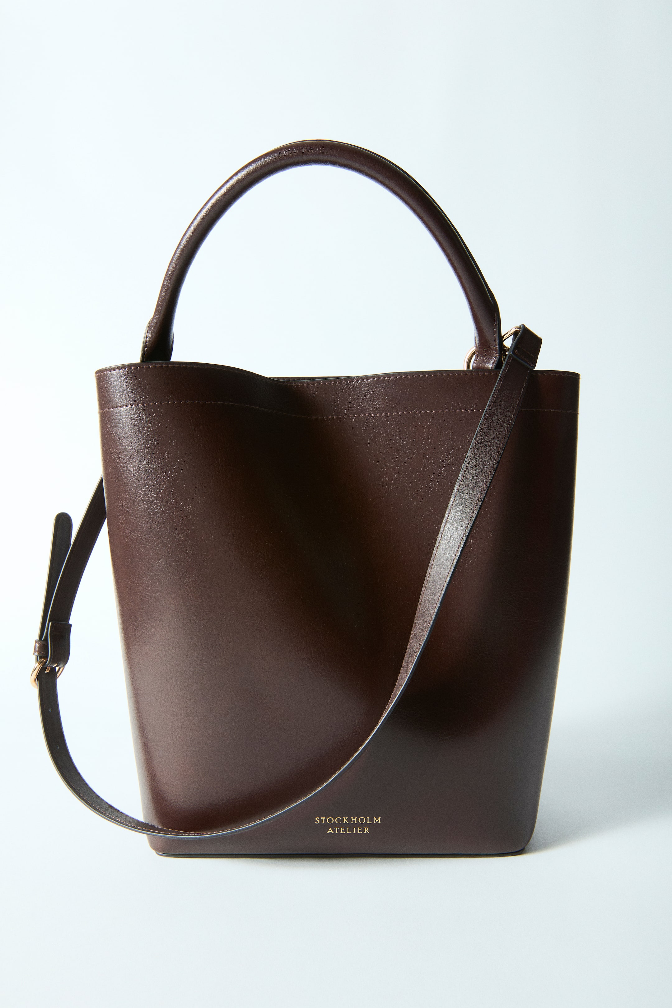 View larger image: A dark brown bucket bag is facing front, featuring a single rounded top handle and a detachable, adjustable shoulder strap. Gold 'STOCKHOLM ATELIER' branding is visible on the bag's lower front.