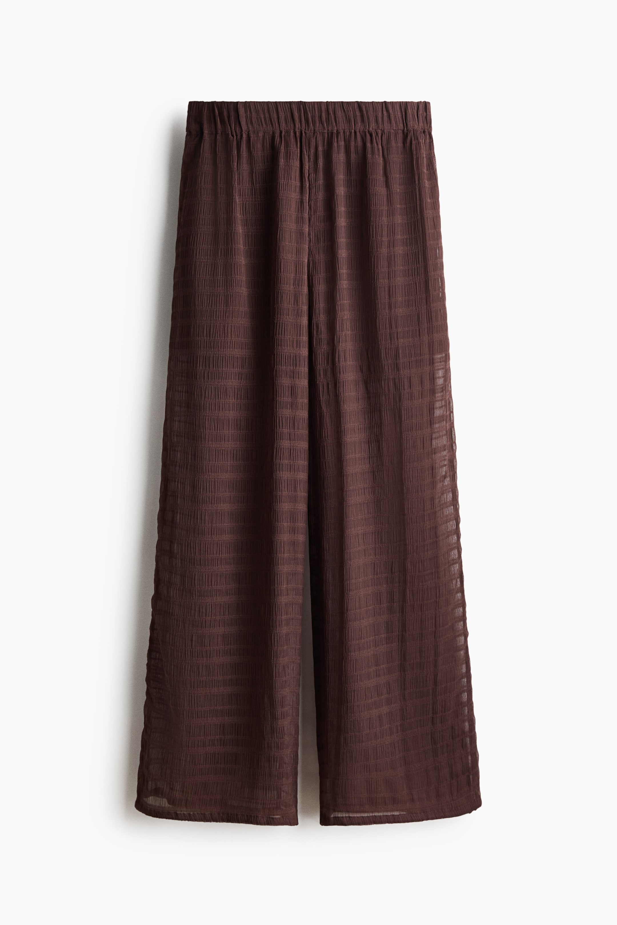View larger image: Dark brown wide-leg trousers are featuring an elasticated, gathered waistband. The sheer fabric is displaying a subtle textured grid pattern and a crinkled effect, forming a relaxed, flowing silhouette.