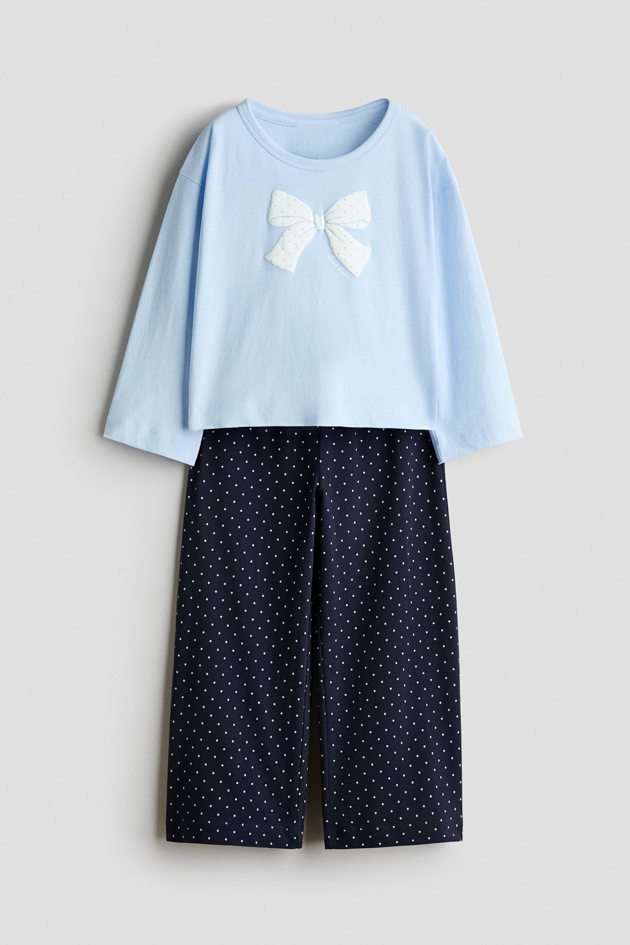 View larger image: A two-piece pajama set is shown facing front, consisting of a light blue long-sleeved top and navy blue straight-leg pants. The top features a crew neck and a white textured bow appliqué on the chest, while the pants display an all-over white polka dot pattern.