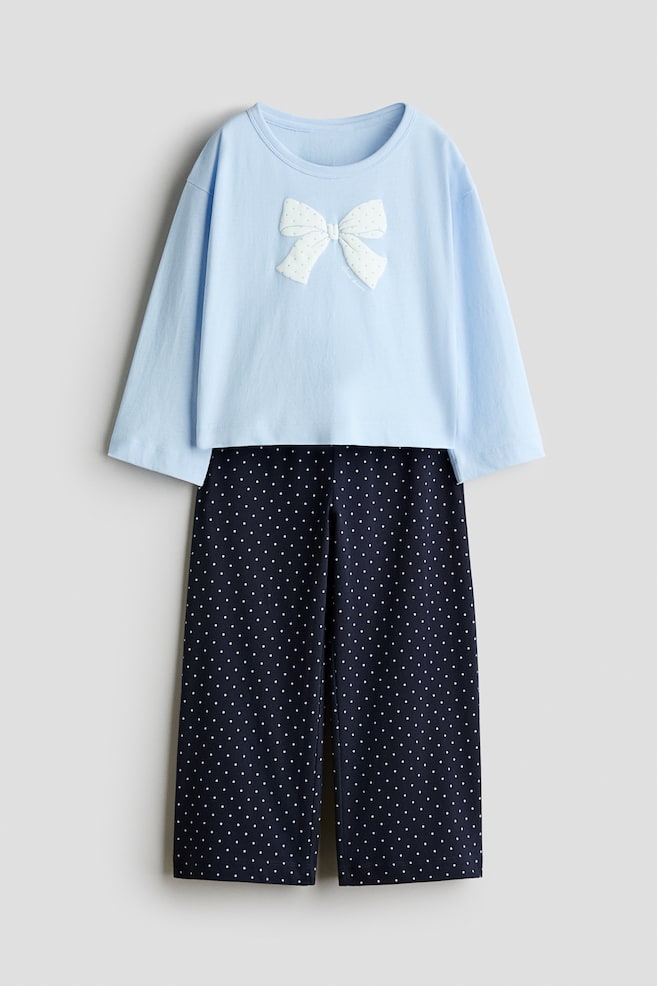 A two-piece pajama set is shown facing front, consisting of a light blue long-sleeved top and navy blue straight-leg pants. The top features a crew neck and a white textured bow appliquÃ© on the chest, while the pants display an all-over white polka dot pattern. - 1