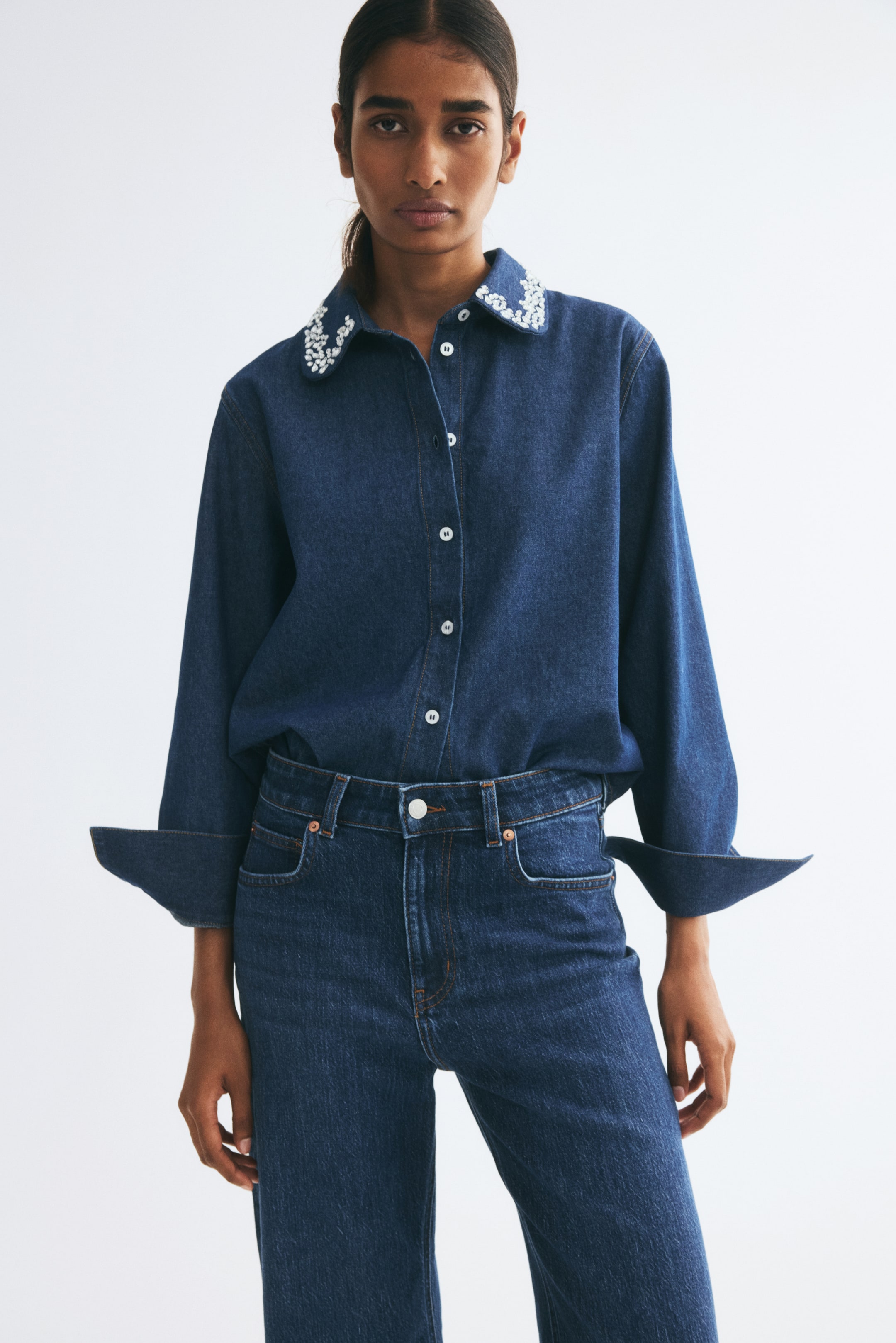 View larger image: A dark blue denim shirt featuring a collar embellished with sparkling white rhinestones is being worn by the model. The shirt is buttoned down the front and tucked into matching high-waisted blue jeans, with its long sleeves styled with wide, turned-up cuffs.
