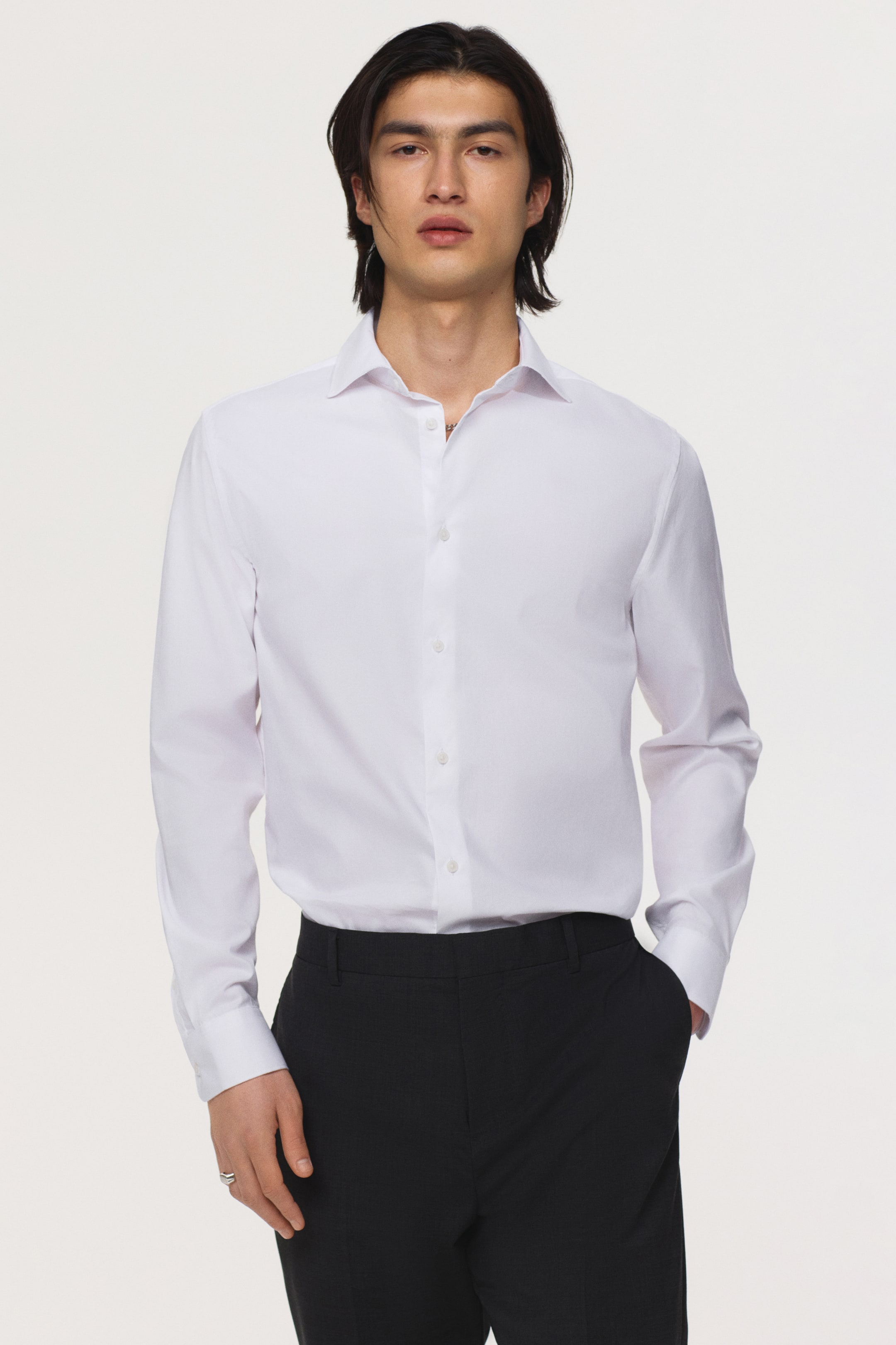 View larger image: A white dress shirt is showcased from the front, featuring a spread collar and long sleeves with single-button cuffs. It displays white buttons along the front placket and is styled with a slim fit, tucked into black trousers.