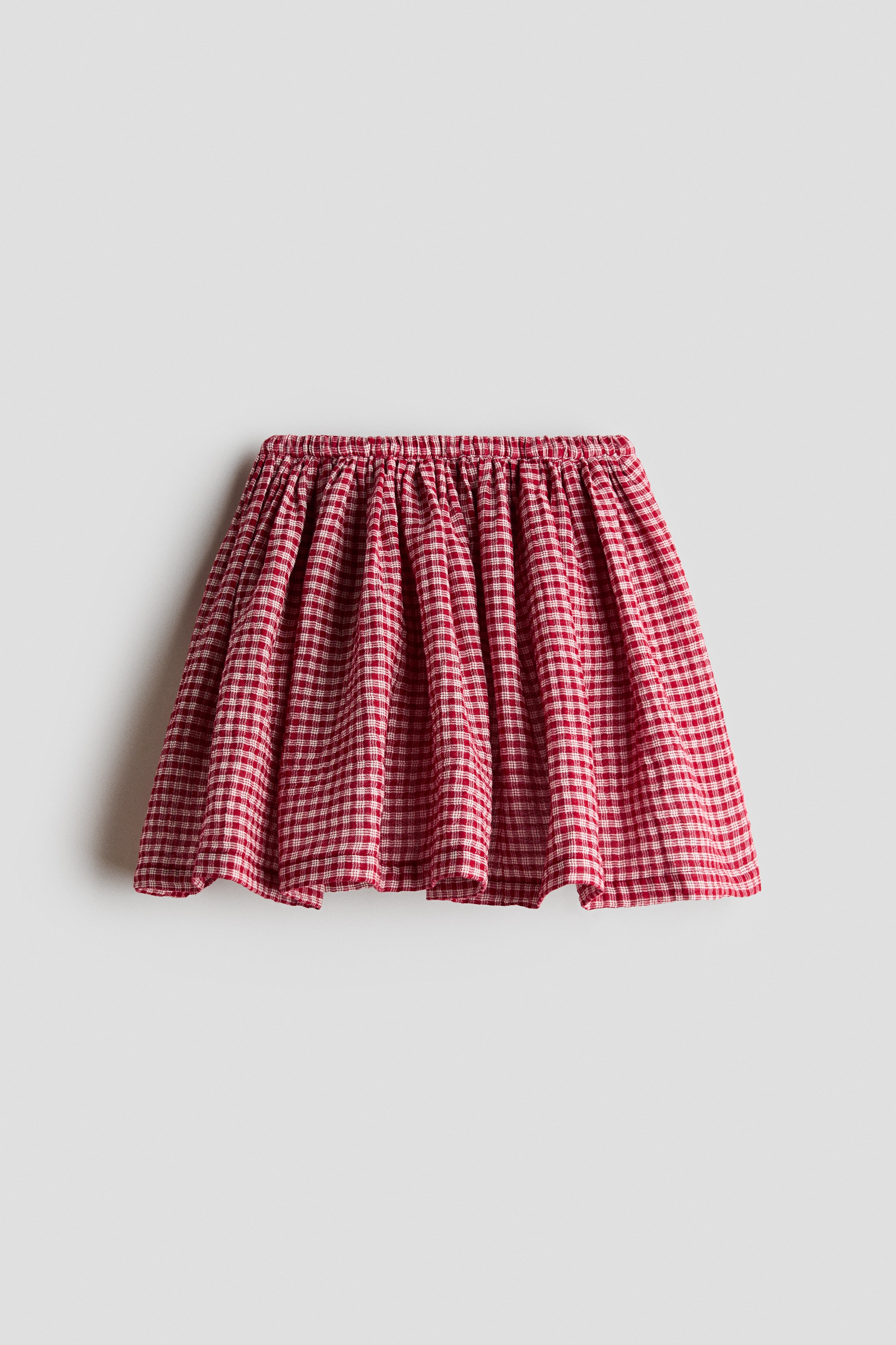 A red and white gingham skirt is shown from the front, featuring a gathered, elasticated waistband that creates a flared, knee-length silhouette. The skirt is paired with a tucked-in white long-sleeved top.