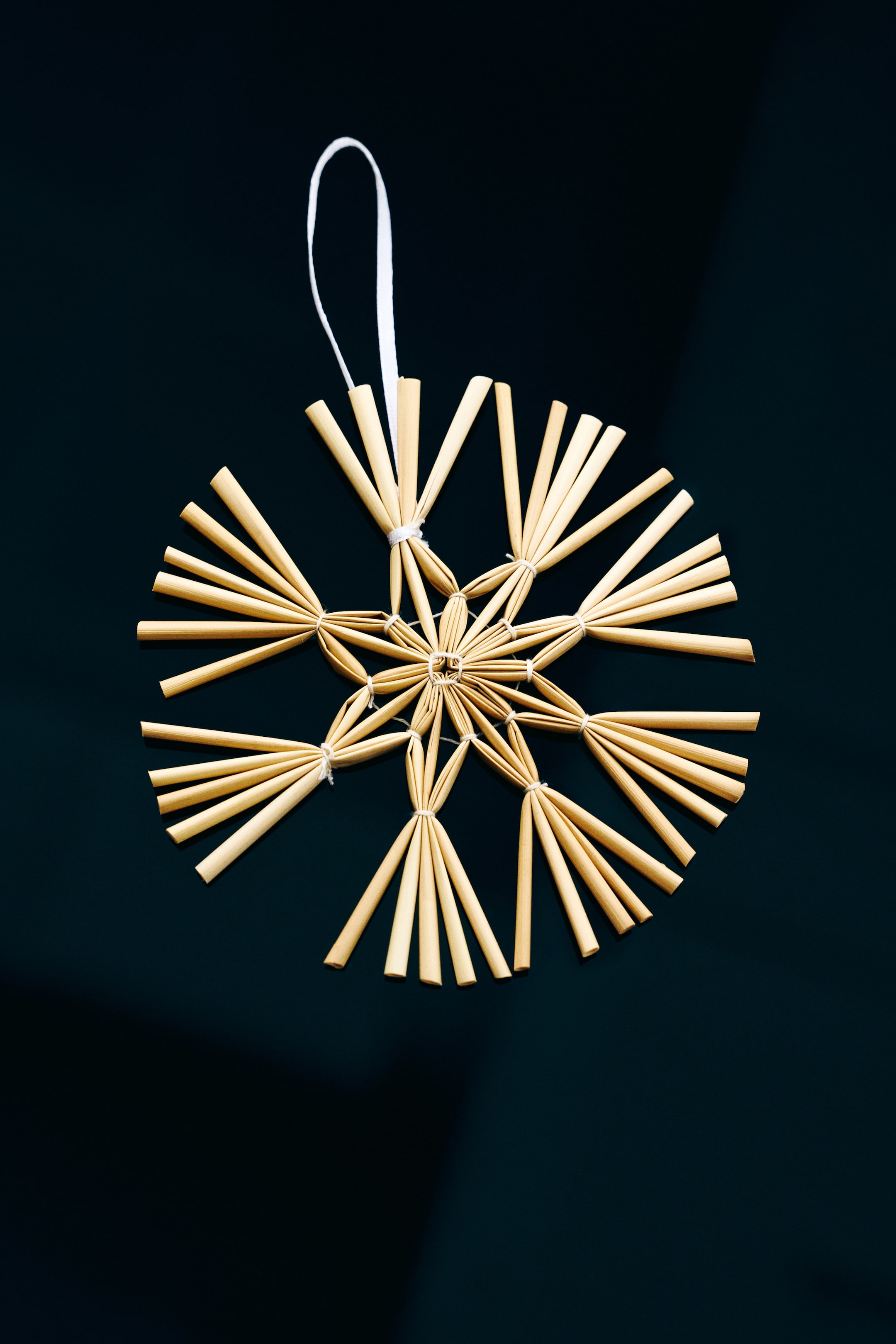 Ver imagem maior: Vista frontal de um ornamento de Natal em forma de estrela, feito de palha natural, apresentando um design geométrico de múltiplas pontas, unido com fio fino, com um laço branco a sair da parte superior para pendurar.