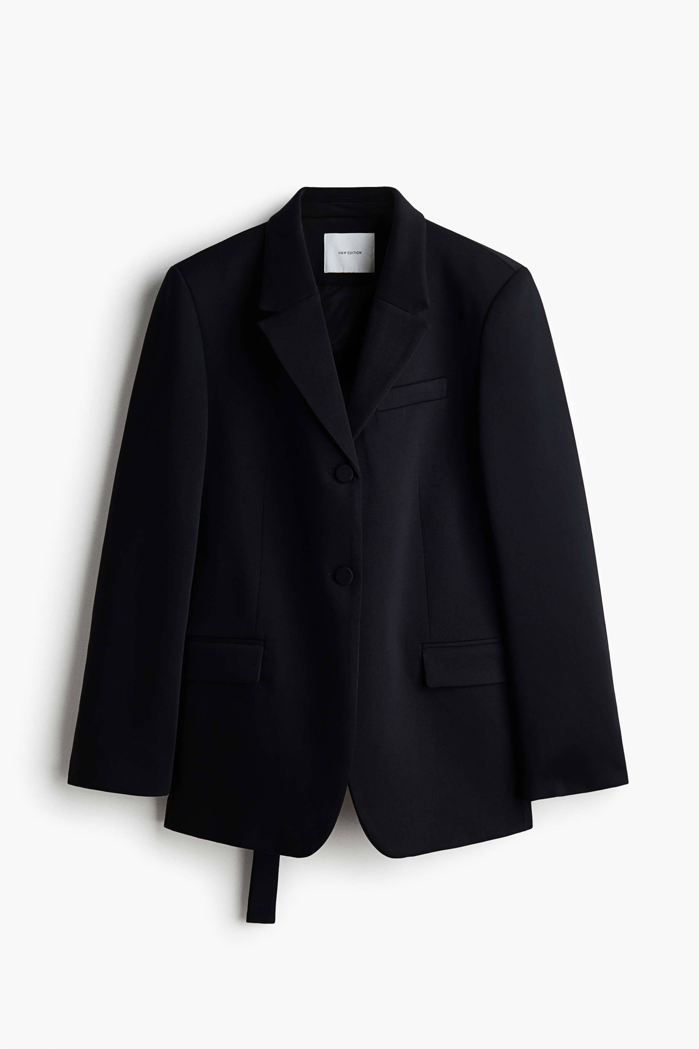 A black oversized blazer with a partially open back is being worn by the model over a cream-coloured shirt with vertical stripes. The back cut-out features a horizontal strap running across the lower back.