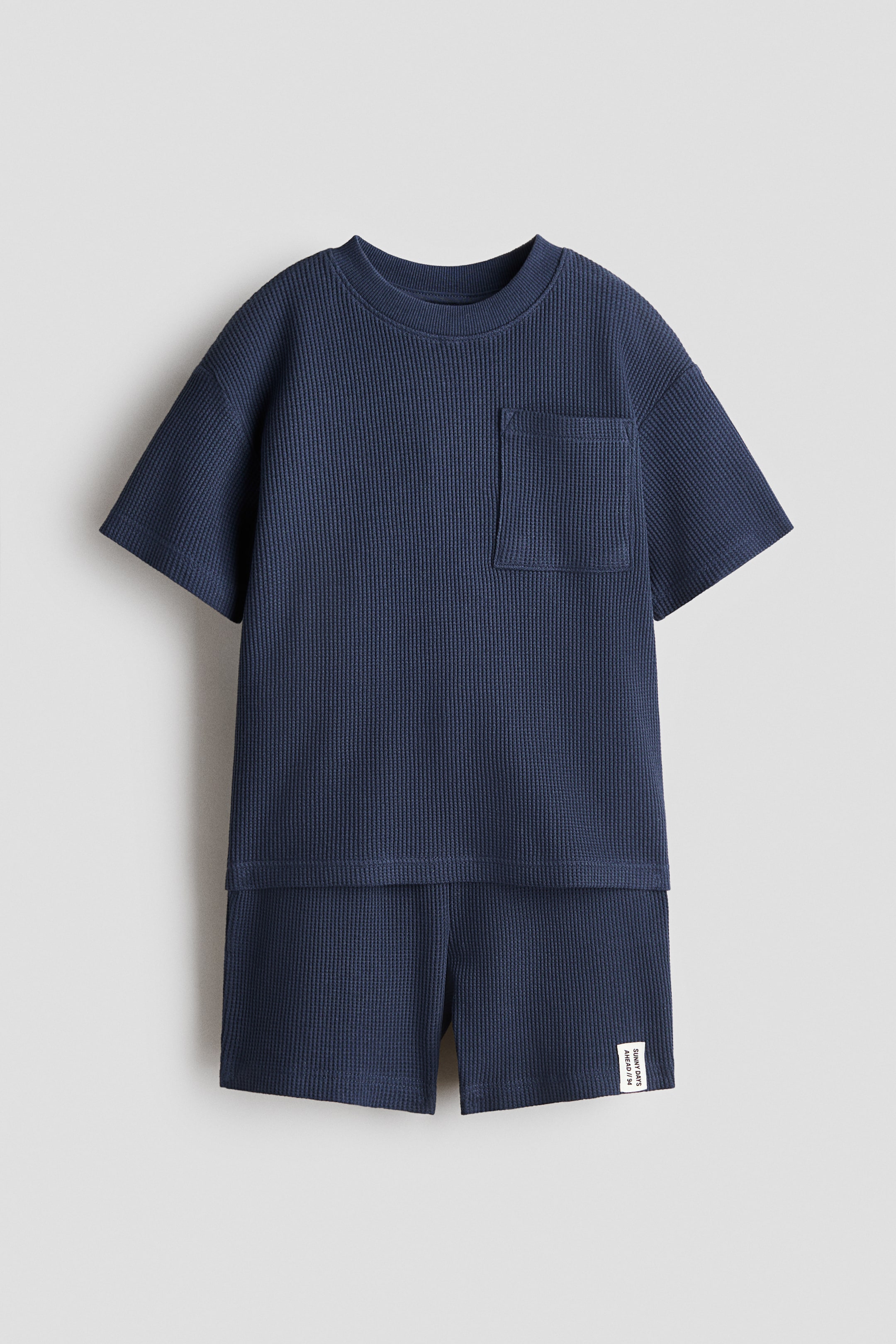 View larger image: A navy blue waffle knit two-piece set is showcasing its front, comprising a t-shirt and shorts. The t-shirt is highlighting a ribbed crew neck, short sleeves, and a chest patch pocket. The matching shorts are displaying a small white fabric label with black text on the right leg.