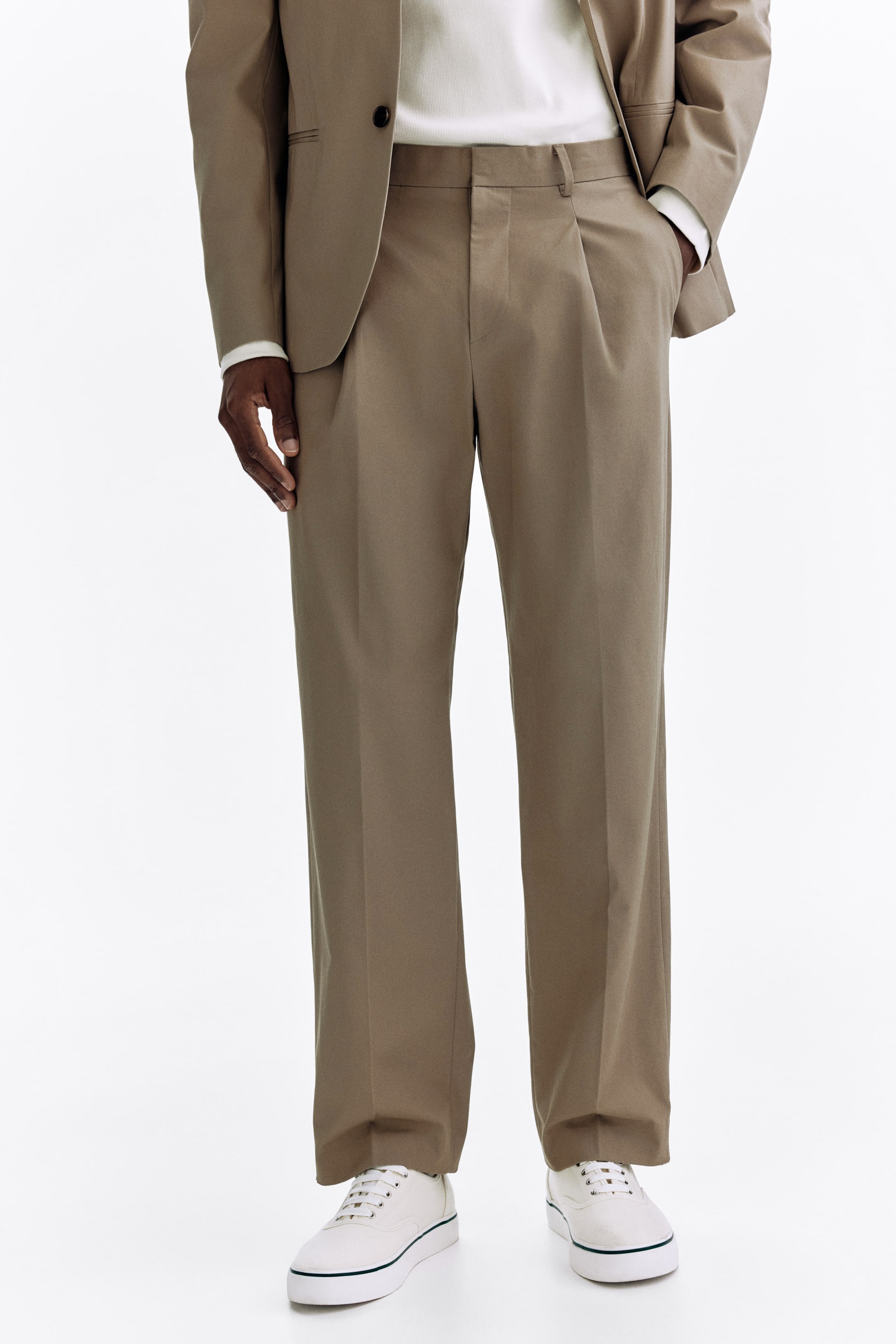 Agrandir l'image: Pantalon plissé marron kaki avec un pli unique sur chaque jambe, une coupe droite et une longueur complète tombant sur des baskets blanches. Le pantalon présente un devant plat avec des passants de ceinture visibles, une fermeture à bouton et des poches latérales.