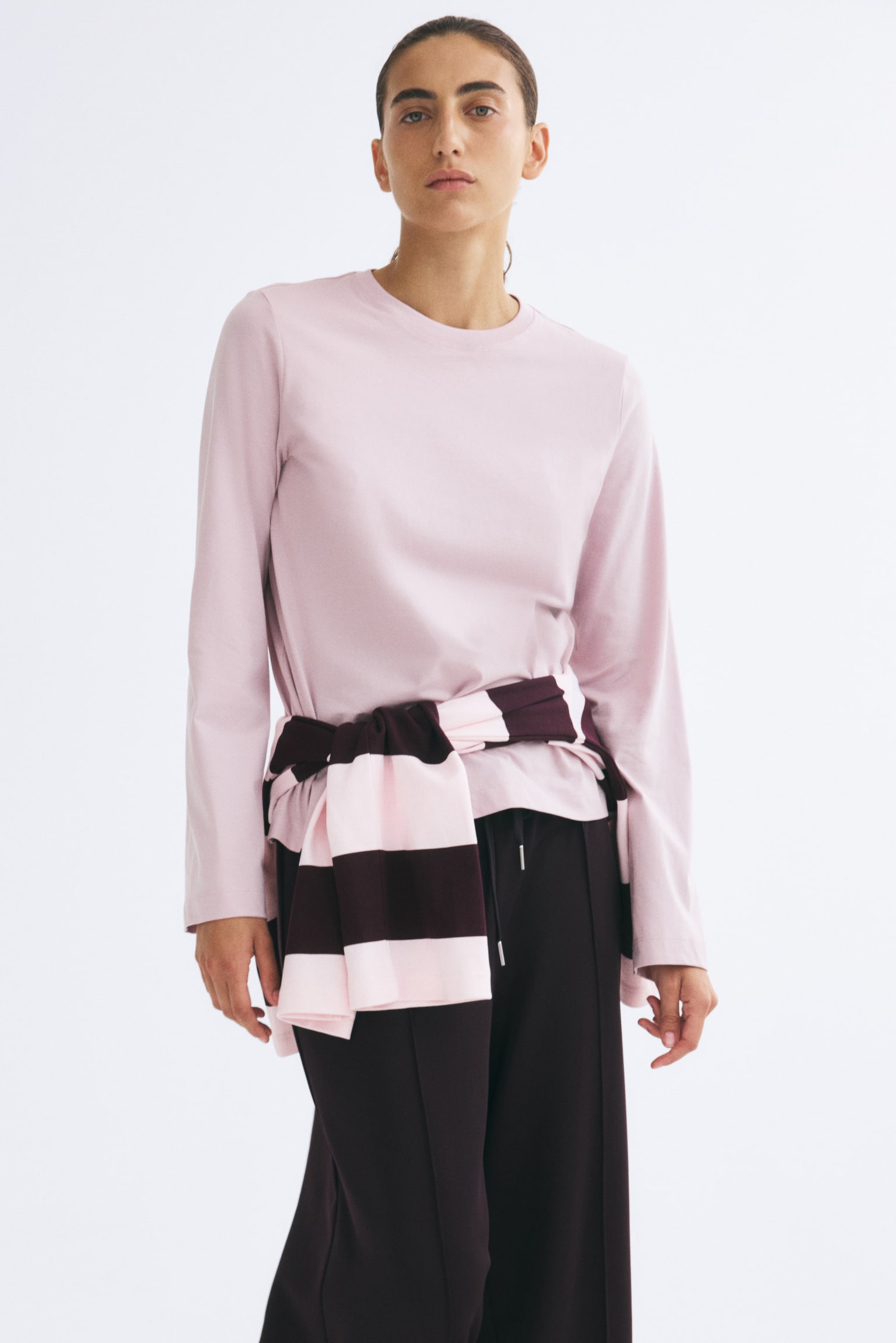 The model is wearing a light pink long-sleeved crew-neck top, styled with a striped pink and dark burgundy sweater tied at the waist. The look is finished with dark, relaxed-fit trousers. - 1