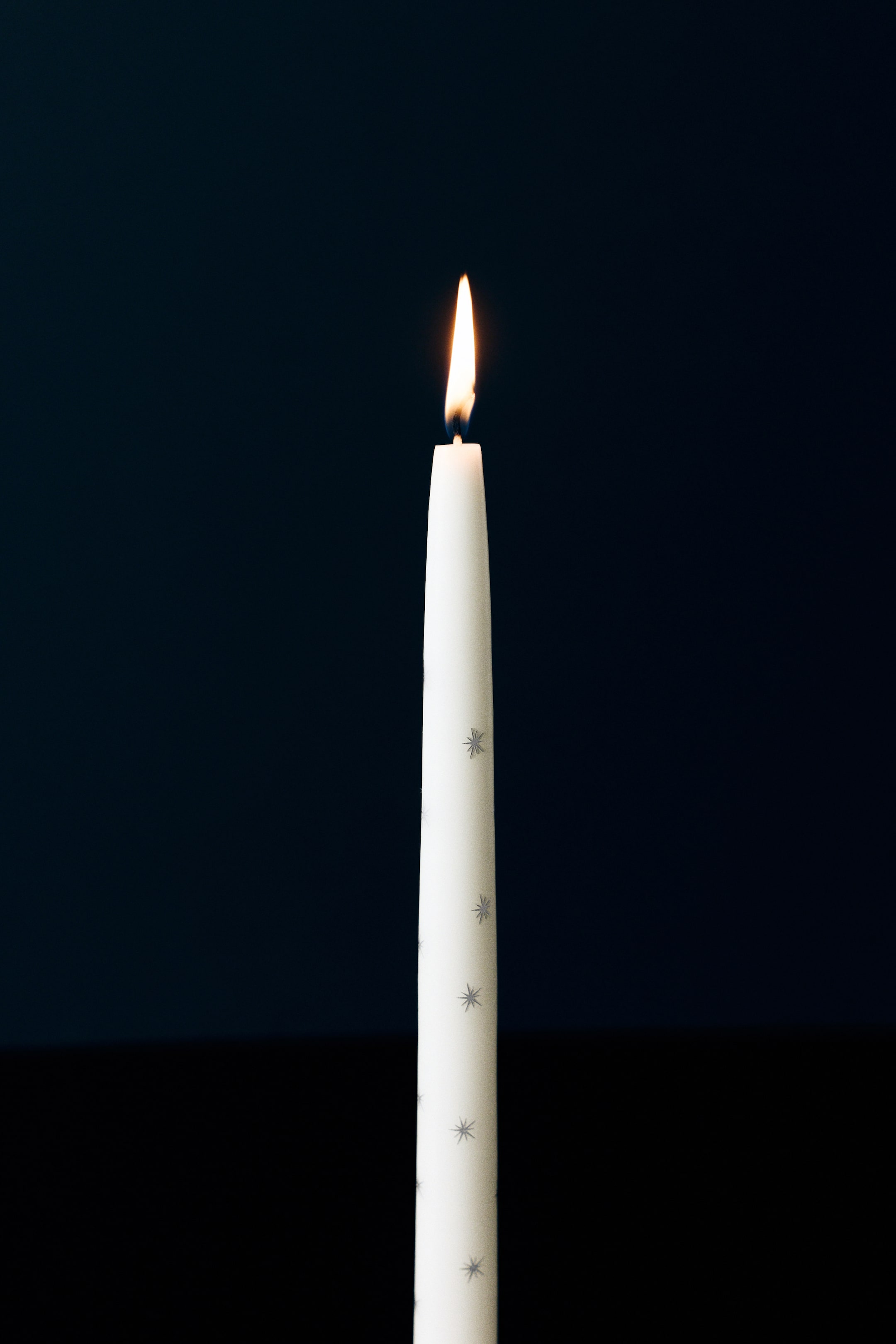 Agrandir l'image: Une bougie conique blanche ornée d’un motif intégral de petites étoiles scintillantes argentées brûle avec une flamme vive à sa mèche.
