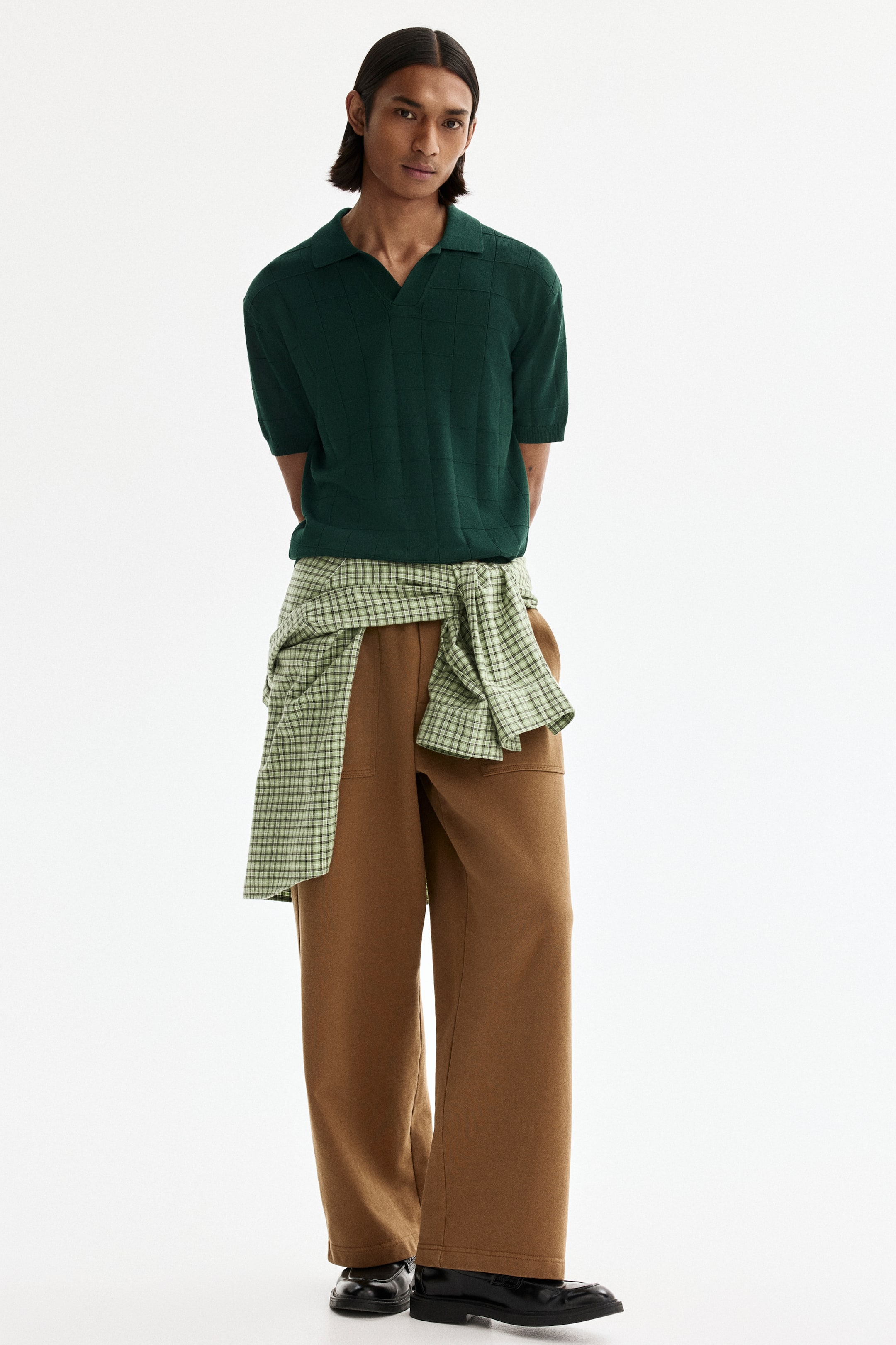 Ampliar la imagen: Se presenta un polo de manga corta de punto color verde oscuro, con un sutil patrón de cuadrícula. Está combinado con pantalones café de pierna ancha que presentan bolsillos parche en la parte delantera; una camisa a cuadros en verde claro y blanco está anudada a la cintura, y en los pies se llevan mocasines negros.
