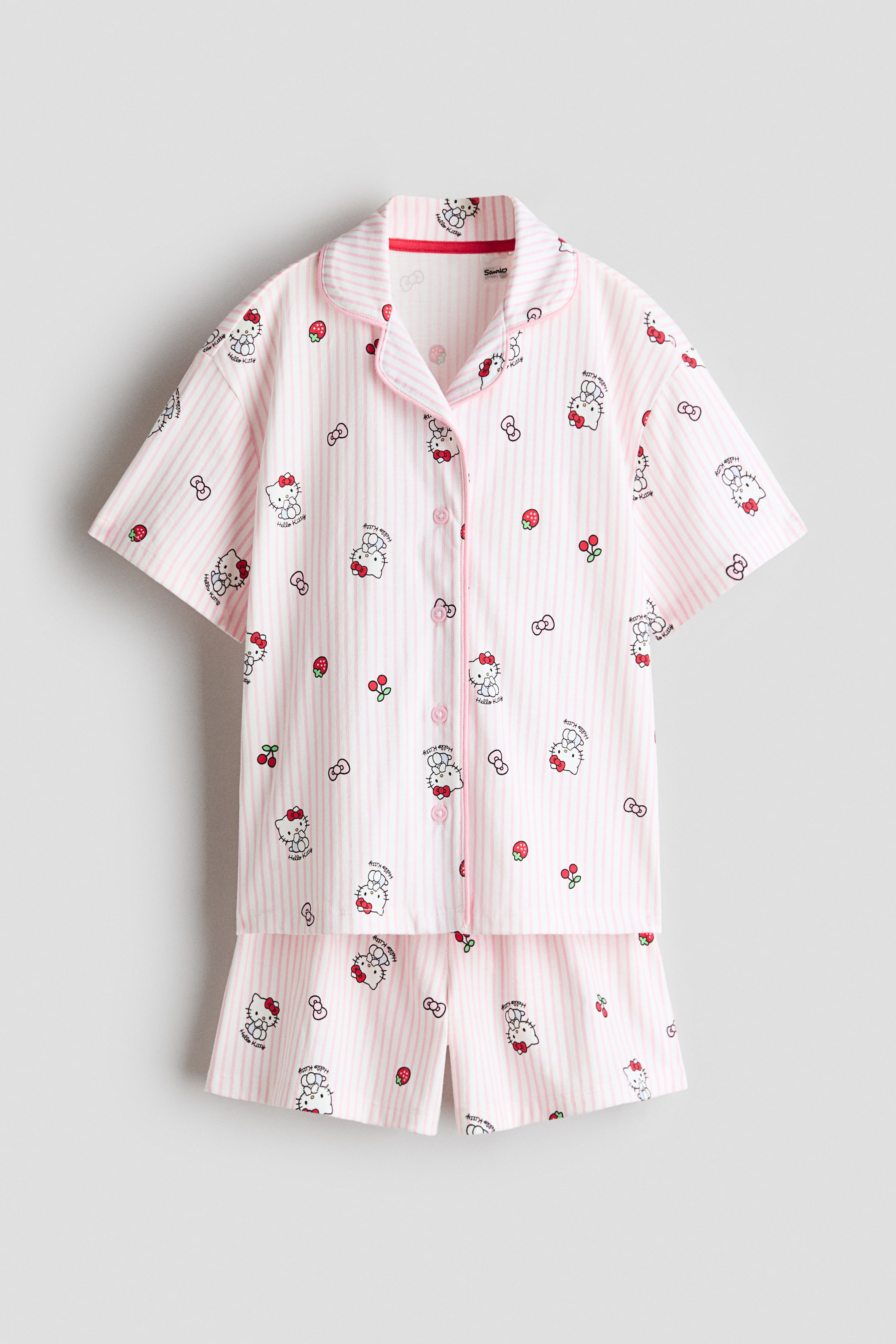 Visualizza immagine più grande: Un set pigiama a maniche corte con pantaloncini coordinati è presentato da una vista frontale diretta, caratterizzato da righe verticali rosa e bianche con un motivo ripetuto di personaggi Hello Kitty, fragole, ciliegie e fiocchi. La parte superiore è progettata con un colletto a revers, una chiusura frontale con bottoni rosa e profili rosa che delineano l'abbottonatura, il colletto e i polsini delle maniche.