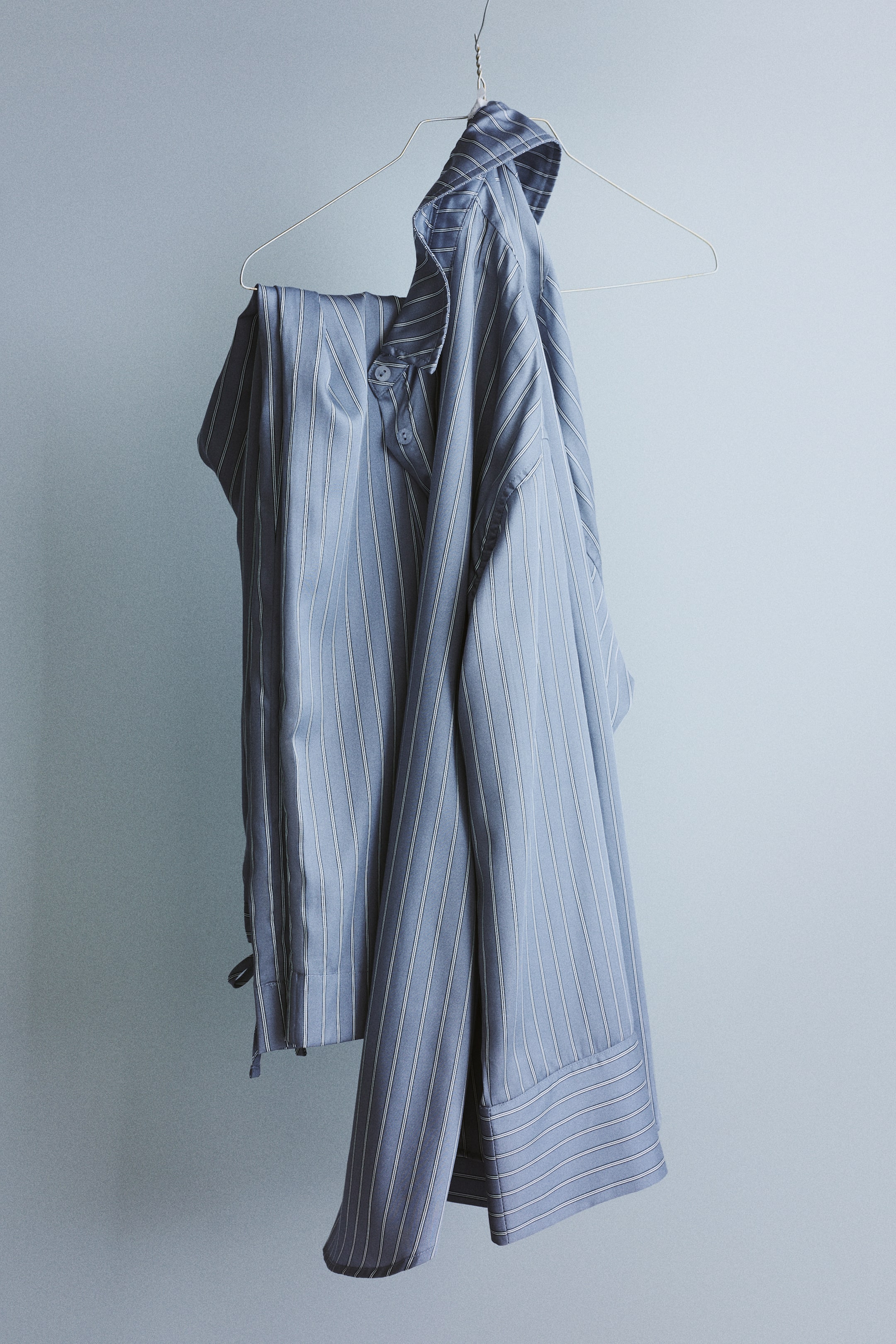 Visa större bild: Pyjamas med en långärmad skjorta och ett par byxor i mjukt fallande lyocell-satin hänger på en galge. Skjortan har ett randigt mönster, nedvikt krage, knappar framtill, lågt nedhasade axlar, vida manschetter och en rundad fåll, medan de matchande randiga byxorna är vikta och hänger nedanför.