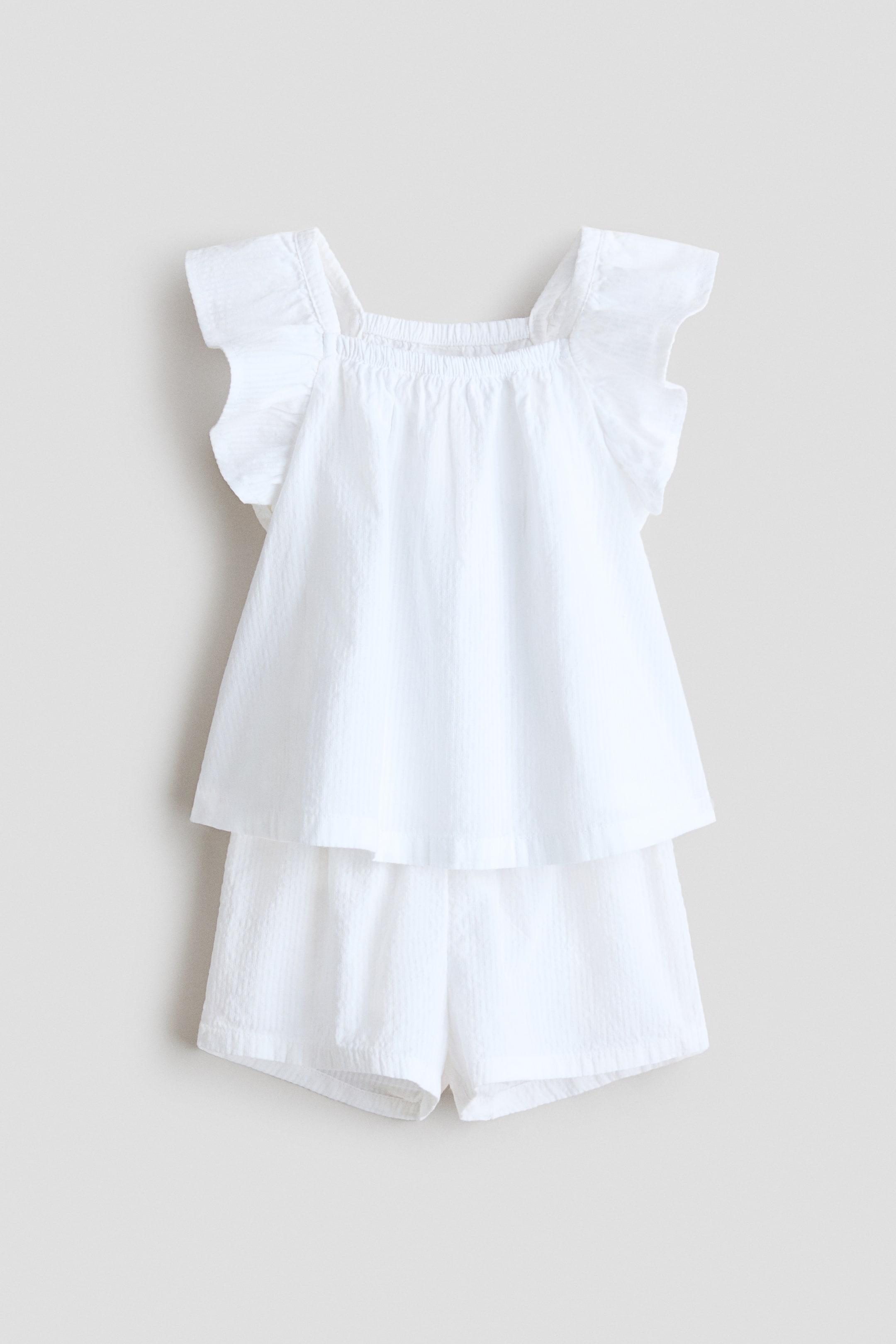 View larger image: A white set, front‑facing, features a top and a pair of shorts in an airy weave. The top has flutter sleeves and a square neckline with covered elastication at the front and back, while the shorts show covered elastication at the waist.