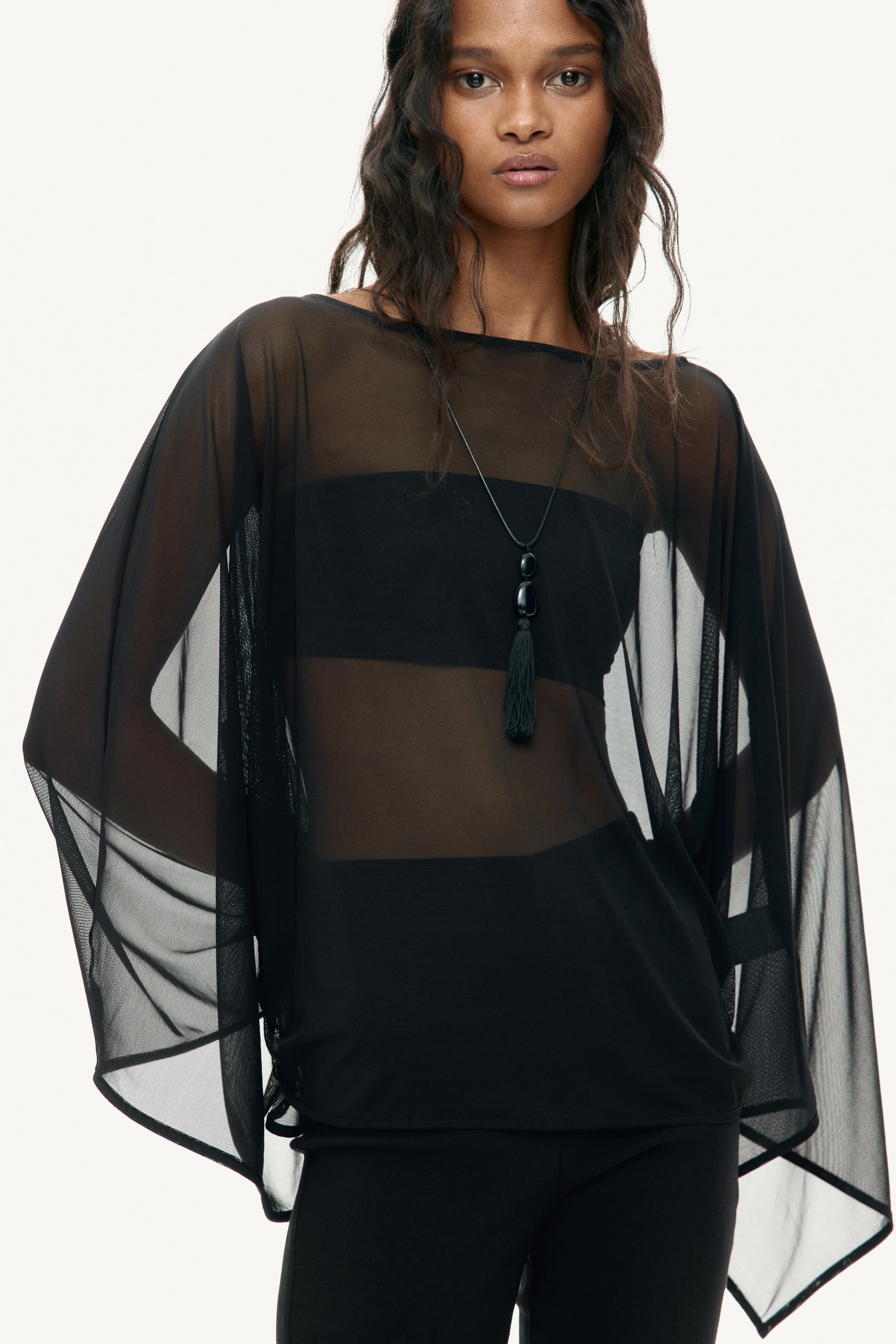 View larger image: A black sheer top featuring a boat neck and wide, flowing sleeves. The transparent, mesh-like fabric reveals a black bandeau top underneath, with a black necklace adorned with two oval beads and a tassel hanging at the front.