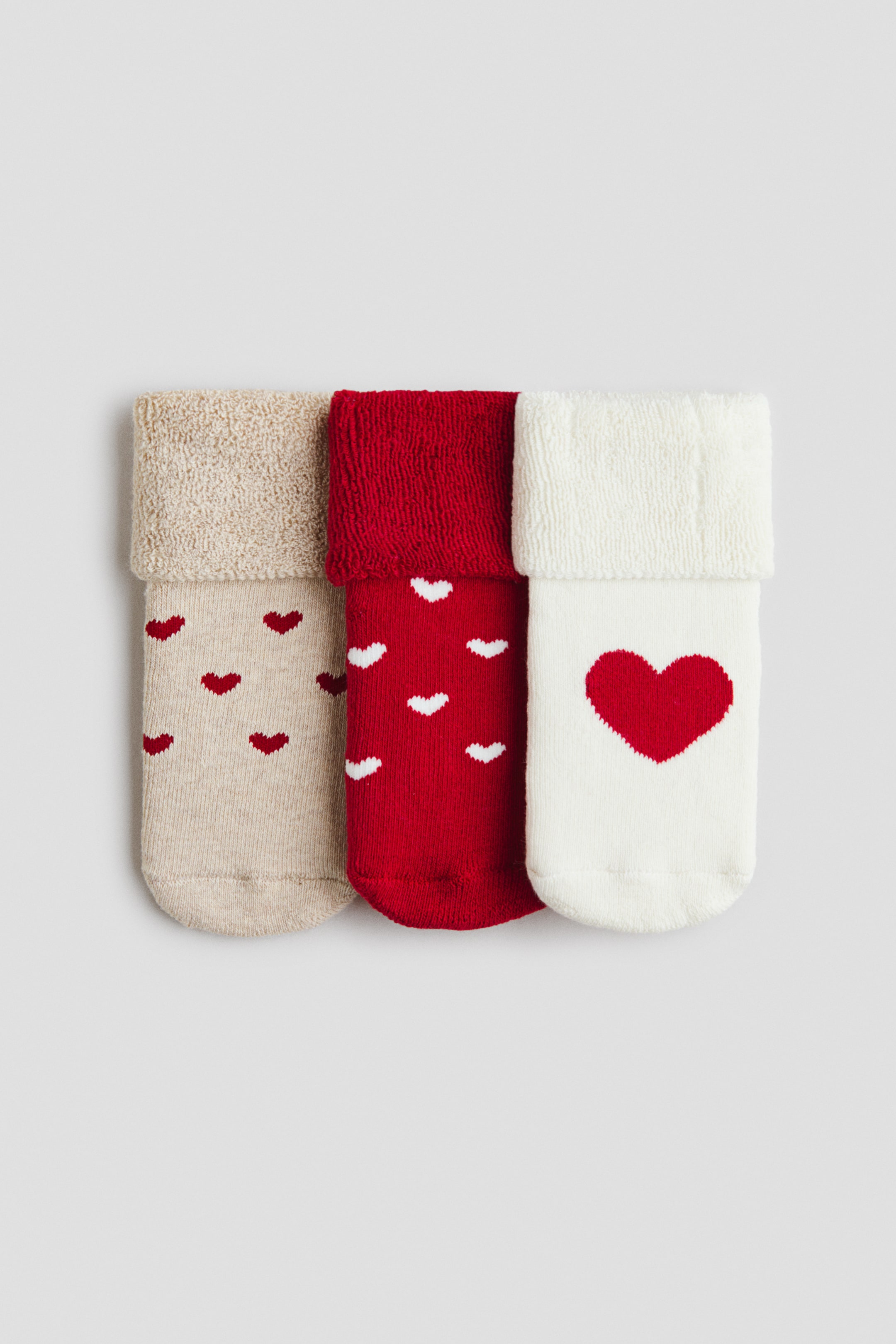 Agrandir l'image: Un ensemble de trois paires de chaussettes en maille fine est présenté de face, chacune dotée d’une tige à revers avec une texture éponge douce. La chaussette de gauche est beige avec de petits cœurs rouges, celle du milieu est rouge avec de petits cœurs blancs, et celle de droite est crème avec un grand cœur rouge.