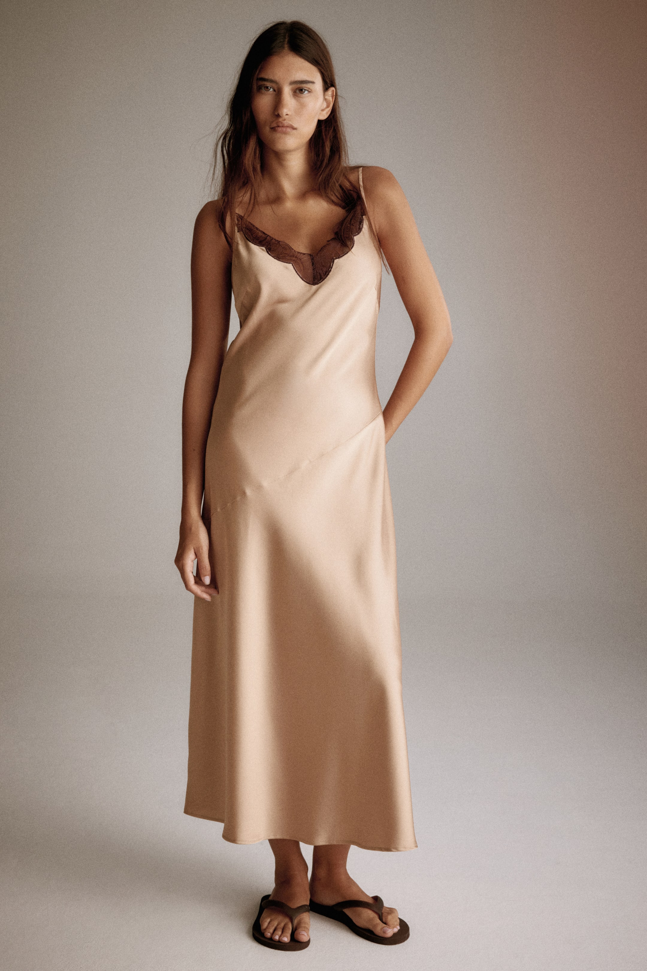 View larger image: A calf-length dress in softly draping satin is being worn by the model, featuring a dark lace-trimmed V-neckline, adjustable spaghetti shoulder straps, a diagonal seam from the waist to the hip, and a gently flared skirt. The model is standing, wearing dark sandals, with one hand resting on the dress at the hip.