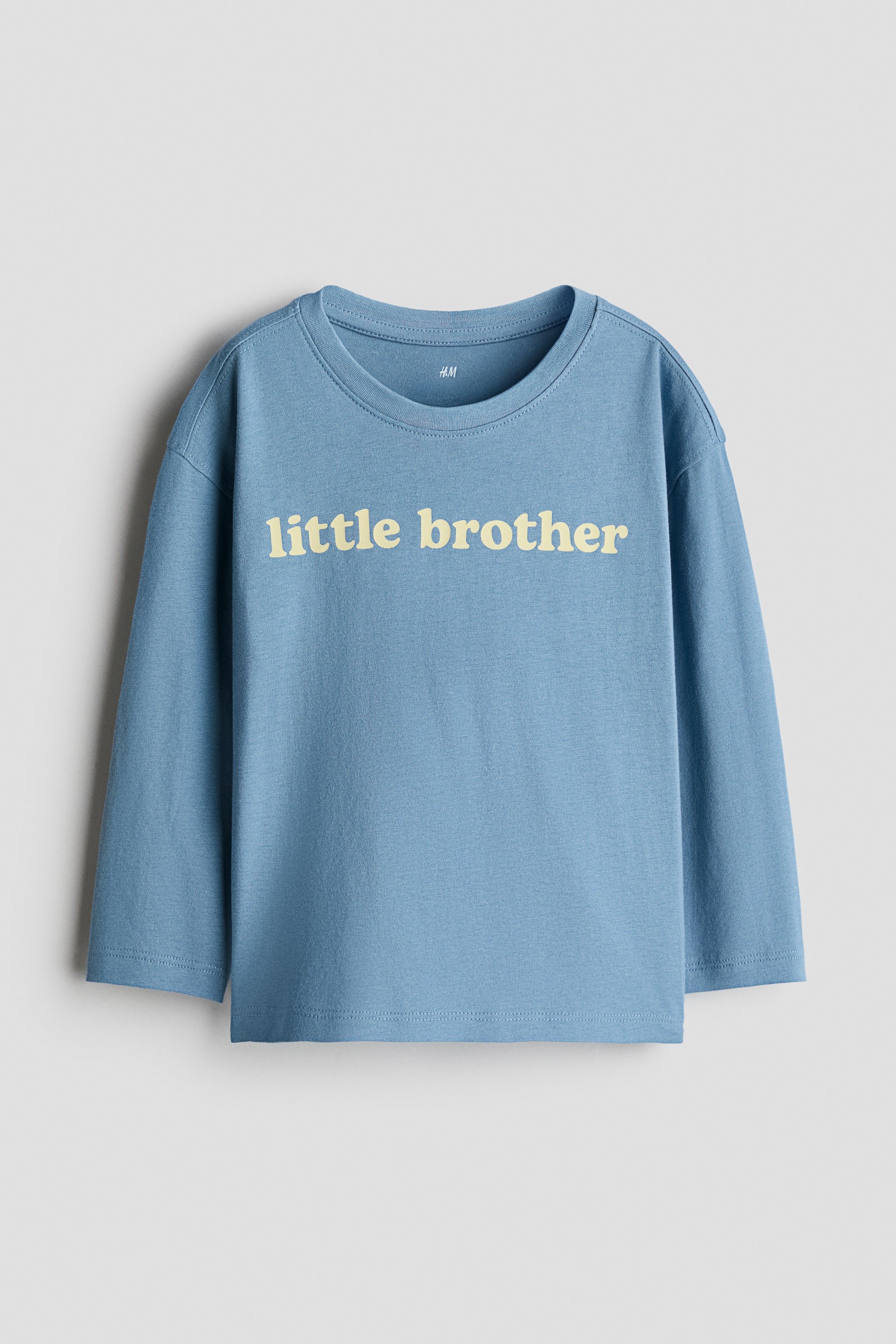 View larger image: A blue long-sleeve cotton jersey T-shirt is shown from the front, featuring a round neckline with ribbed trim and dropped shoulders. The T-shirt displays the text "little brother" printed in a light yellow font across the chest.
