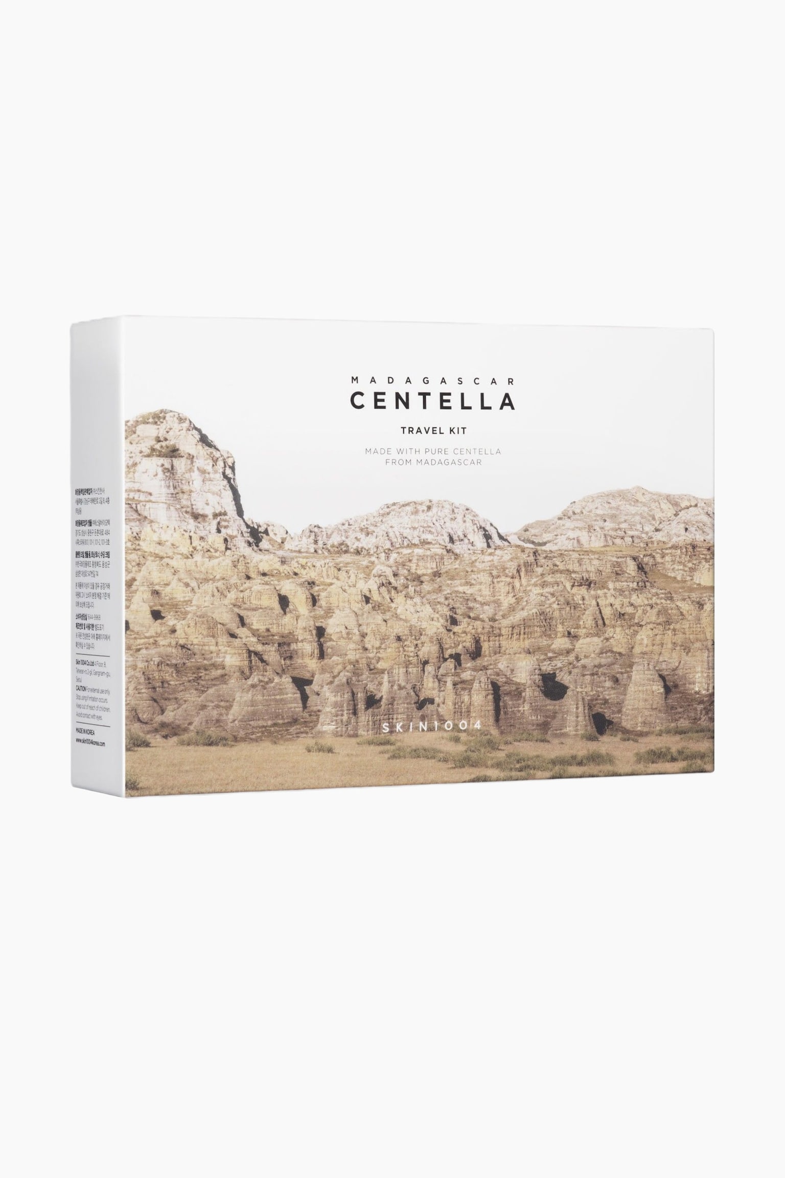 Suorakaiteen muotoinen valkoinen SKIN1004 Madagascar Centella Travel Kit -pakkauslaatikko seisoo kolmen neljäsosan kulmassa oikealle päin. Etupaneelissa on maisemakuva kivikkoisista, beigenvärisistä vuorista, ja brändi sekä tuotteen nimi on painettu mustalla kuvan yläpuolelle. Näkyvissä oleva vasen sivupaneeli näyttää tuotetiedot ja käyttöohjeet mustalla tekstillä.