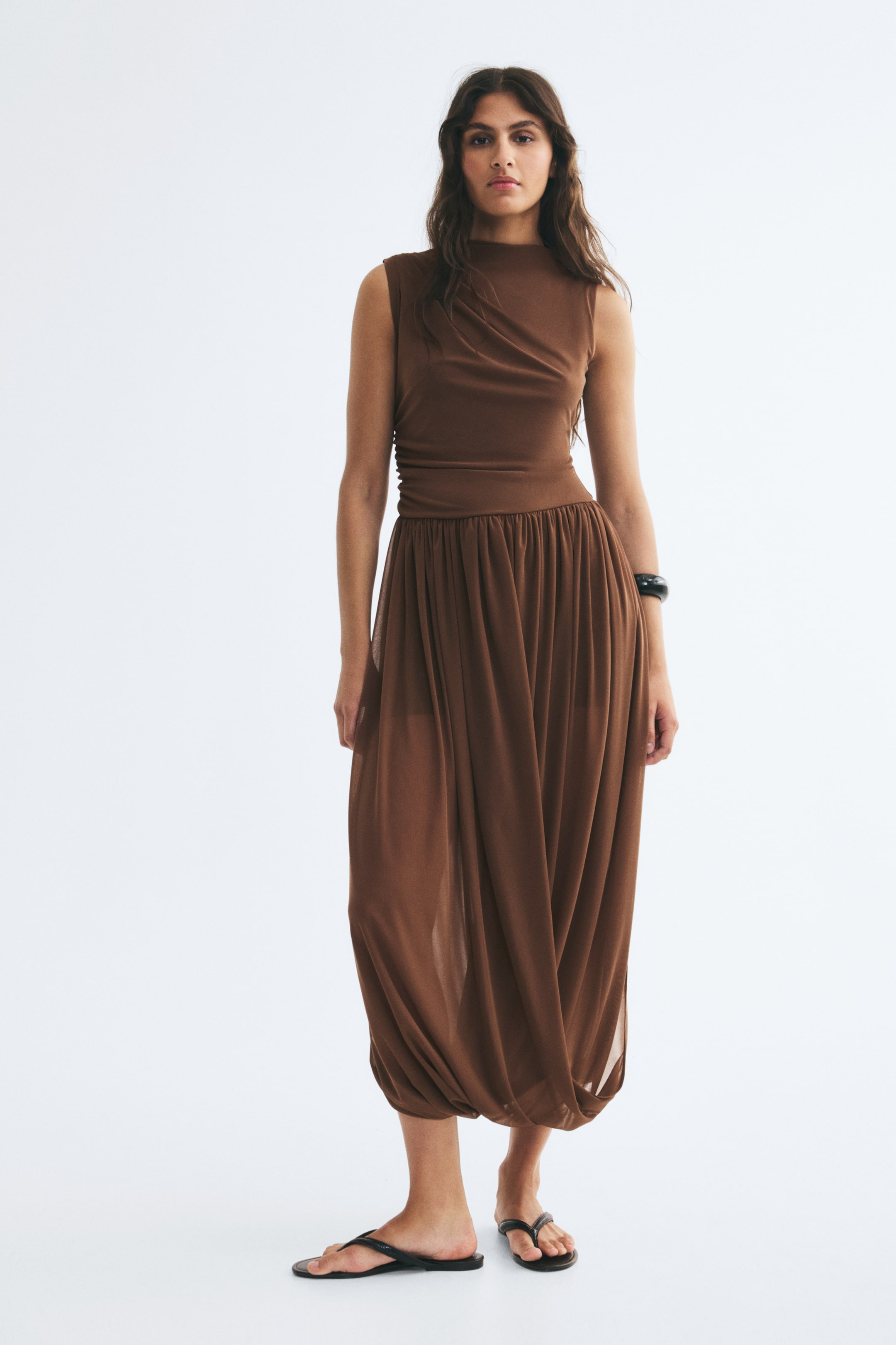 View larger image: A brown, sleeveless, ankle-length dress is being worn by the model, featuring a small stand collar and asymmetrical gathers at the shoulder and waist that create a draped bodice. The dress is styled with a gathered waist and a voluminous, draped skirt.