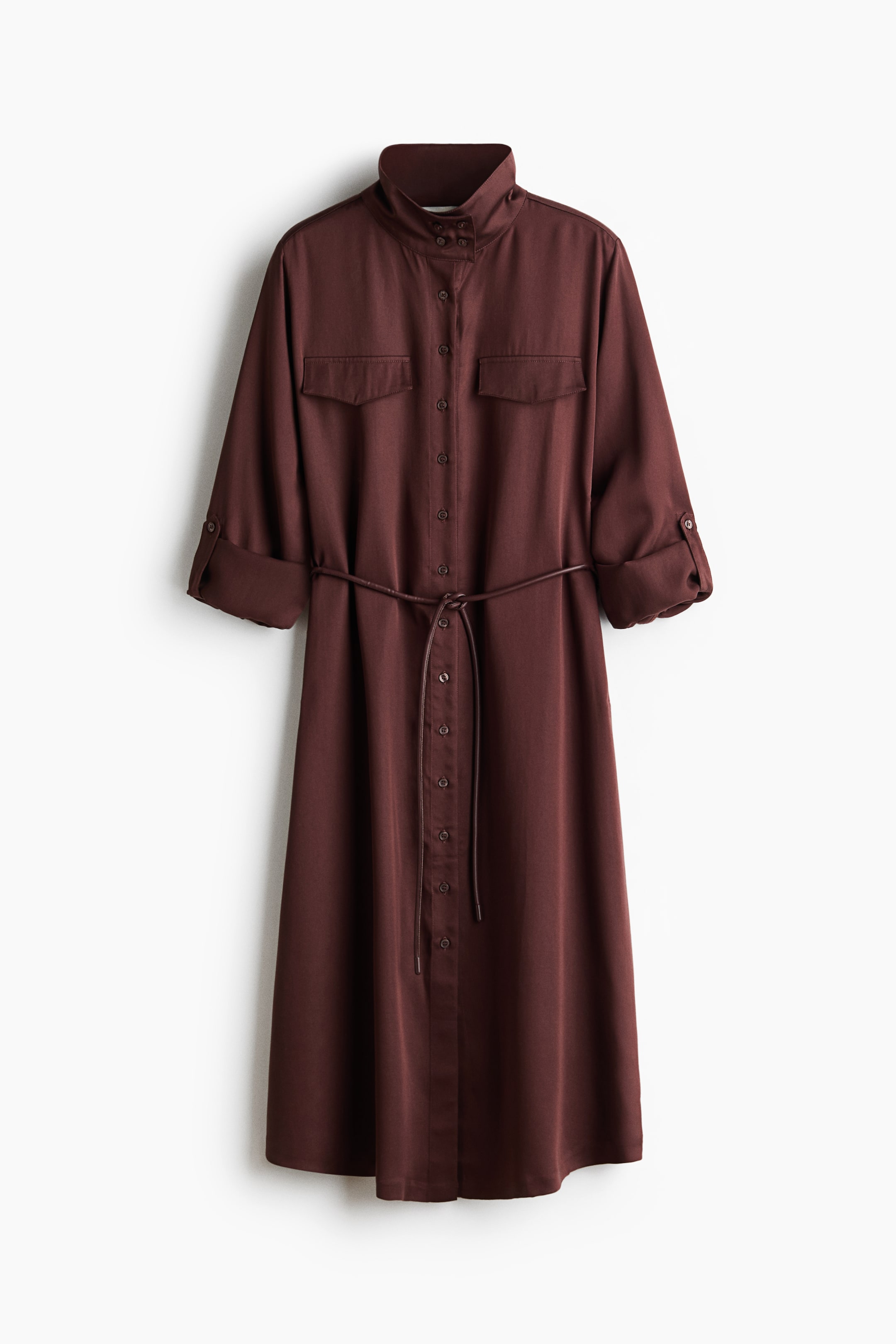 View larger image: A brown shirt dress is hanging straight, featuring a stand-up collar, a full button-down front, and two flap pockets on the chest. The long sleeves are rolled up to a three-quarter length and secured by buttoned tabs, with a thin, matching cord belt cinching the waist of the midi-length dress.