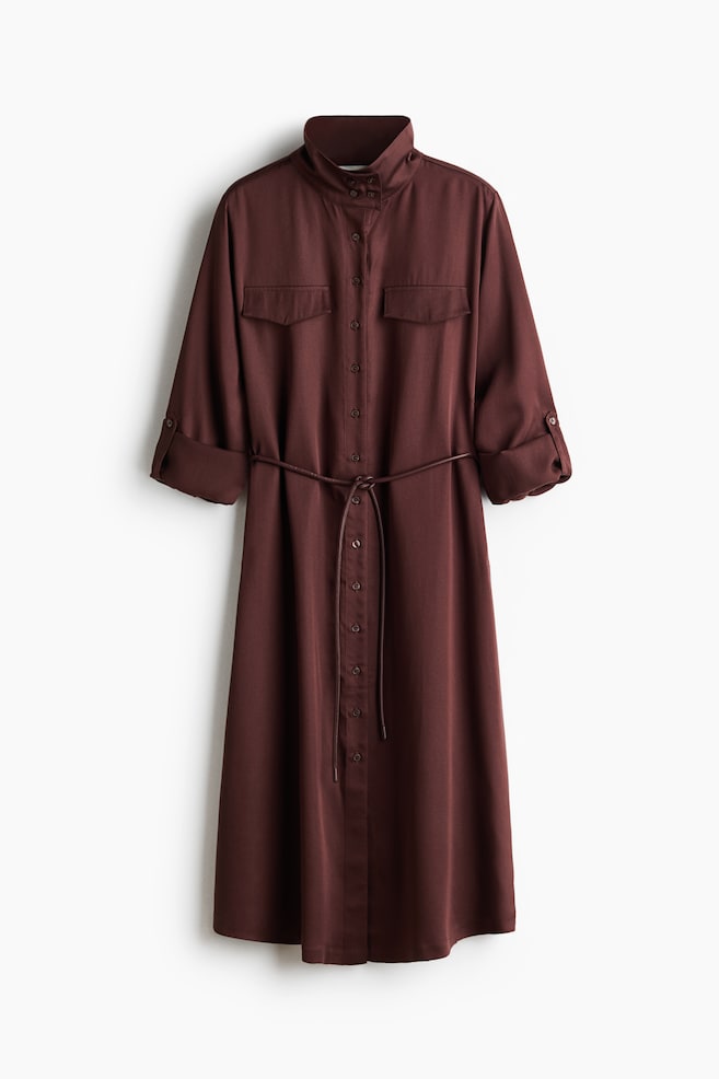 A brown shirt dress is hanging straight, featuring a stand-up collar, a full button-through front, and two flap pockets on the chest. The long sleeves are rolled up to a three-quarter length and secured by buttoned tabs, with a thin, matching cord belt cinching the waist of the midi-length dress. - 1