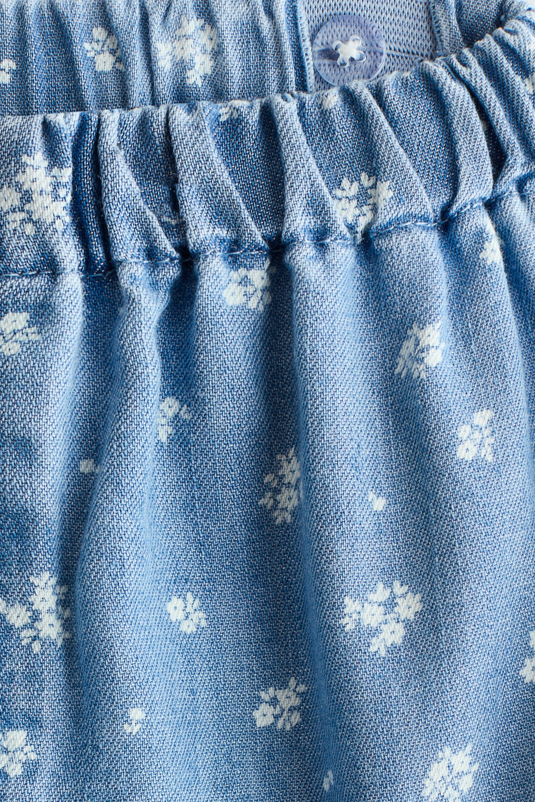 View larger image: This light blue fabric garment showcases a white floral pattern. Its elastic waistline creates soft gathers across the upper front, and a translucent button with a white animal design is visible on the waistband.