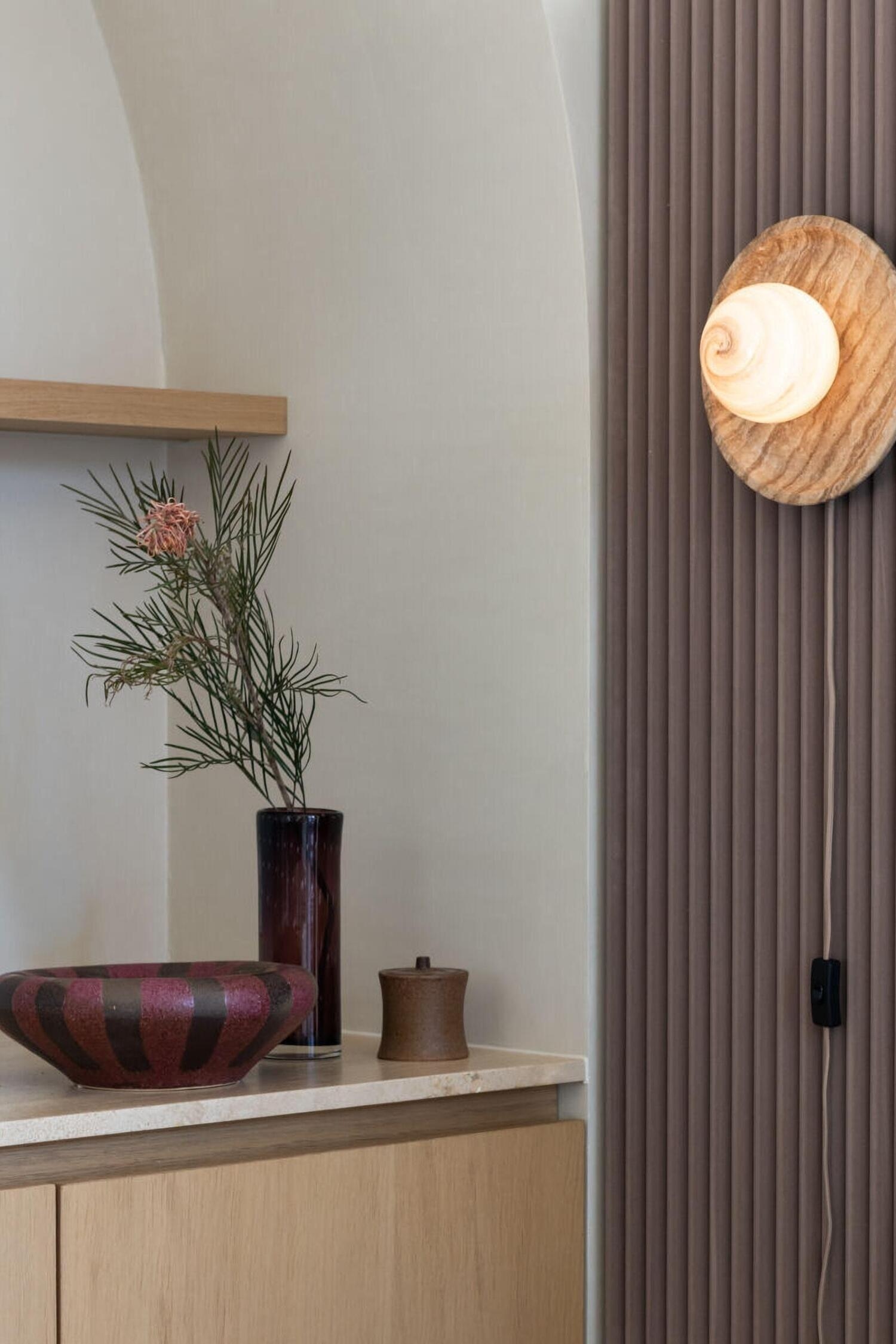 Van opzij gezien toont deze wandlamp een gestructureerde, ronde stenen achterplaat die een verlichte bol ondersteunt. De bol straalt een warme, gemarmerde gele gloed uit, en een bruine stoffen snoer hangt aan de onderzijde.