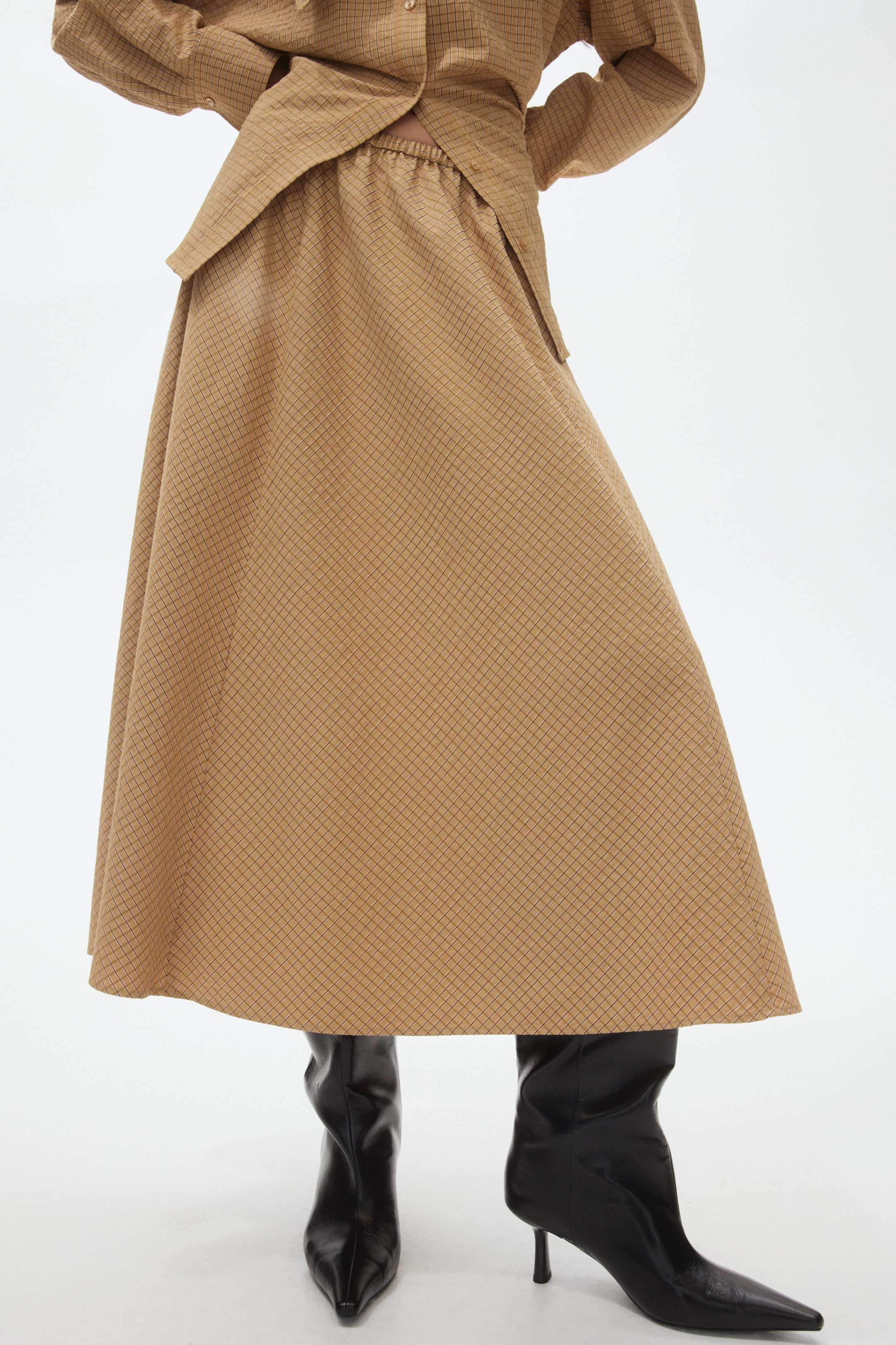 View larger image: A tan midi skirt featuring a subtle grid pattern across its flowing A-line silhouette. It presents an elasticated waistband, partially covered by a matching top, with its hemline falling above black pointed-toe boots.