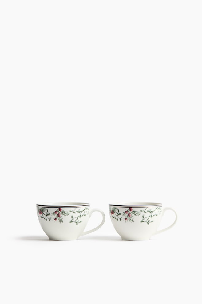 Two white ceramic teacups are sitting side by side, facing forward. Each cup displays a delicate pattern of green pine branches, holly leaves, and red berries along the upper rim, bordered by a thin dark line and featuring a curved handle on the side. - 1