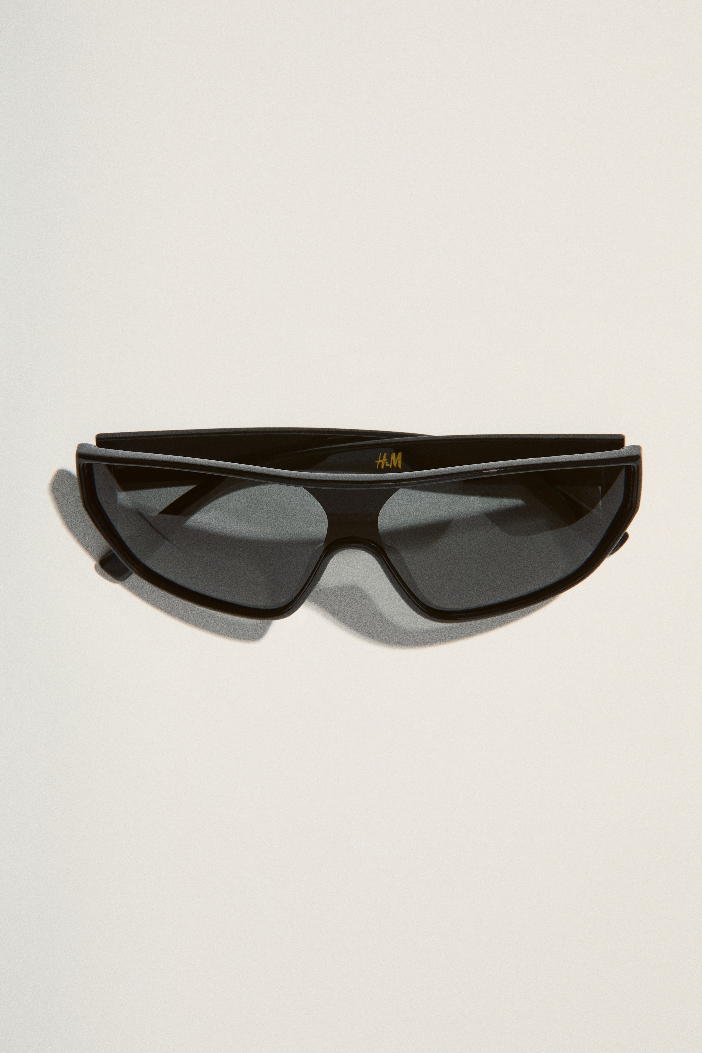 Schwarze Sonnenbrillen im Schildstil liegen flach und präsentieren einen dicken, glänzenden Rahmen mit dunklen Gläsern. Ein kleines 'H&M'-Logo ist auf dem rechten Glas sichtbar, und ein kreisförmiges metallisches Detail ist am linken Bügel zu sehen.