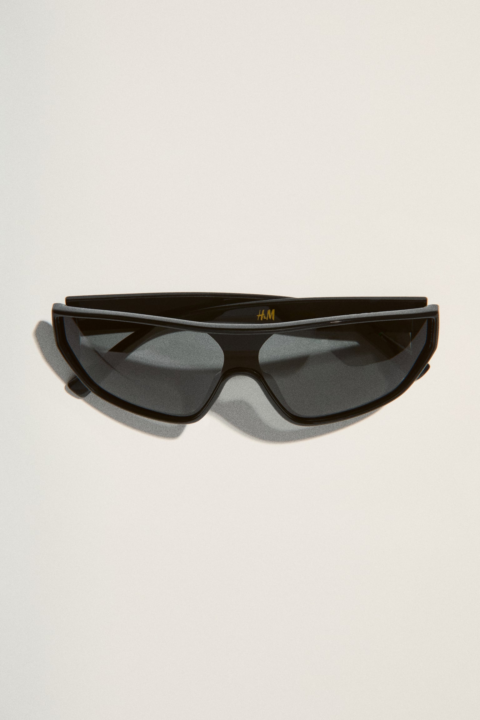 Black shield-style sunglasses are lying flat, showcasing a thick, glossy frame with dark lenses. A small 'H&M' logo is visible on the right lens, and a circular metallic detail is visible on the left temple. - 1