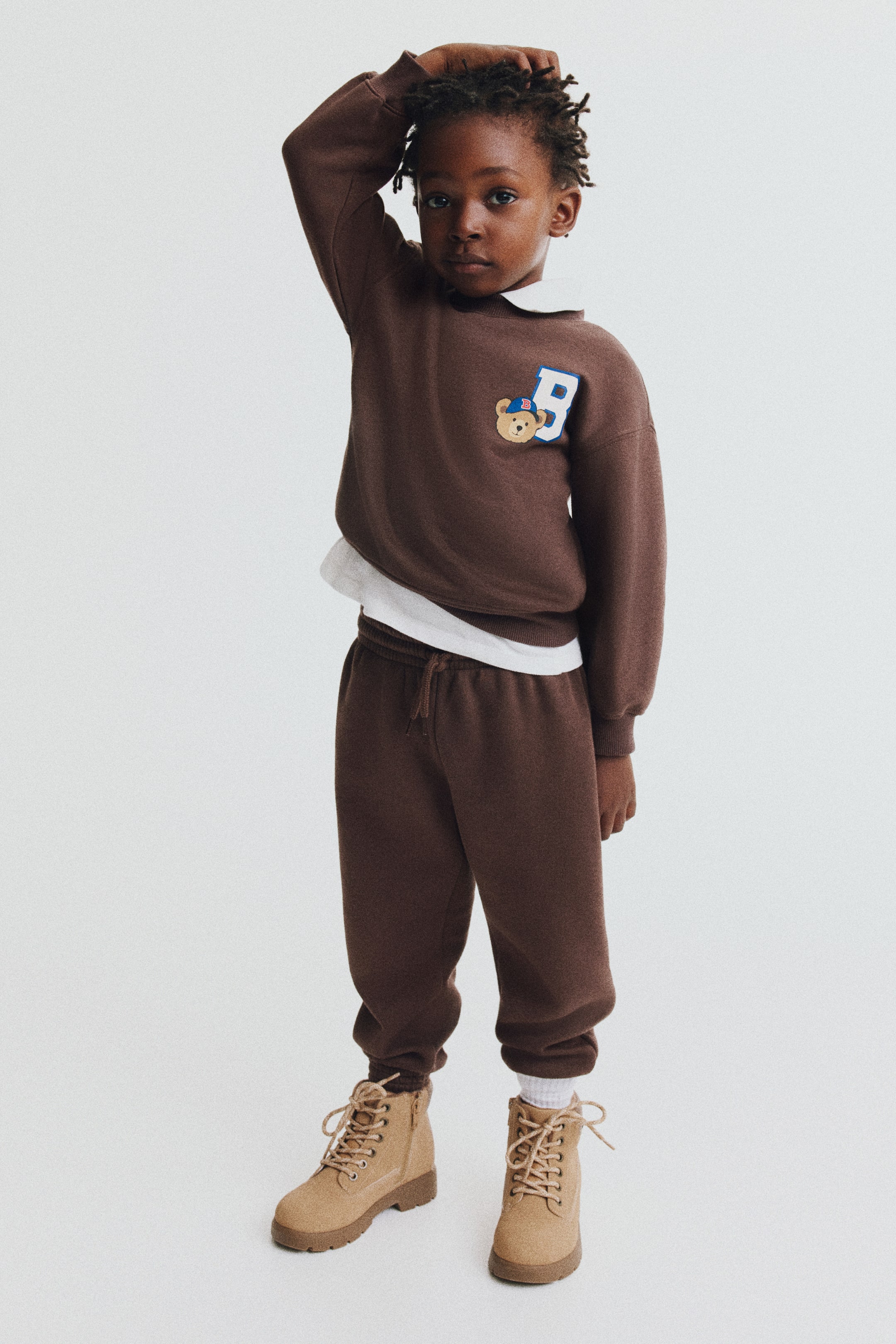 View larger image: This brown sweat-fabric set features a long-sleeved crewneck sweater and matching track pants in a front-facing view. The sweater displays a graphic print of a teddy bear next to a blue letter "B" on the chest, designed with dropped shoulders and ribbed trims, and is layered over a white collared shirt. The matching track pants feature an elasticated drawstring waist and elasticated hems.