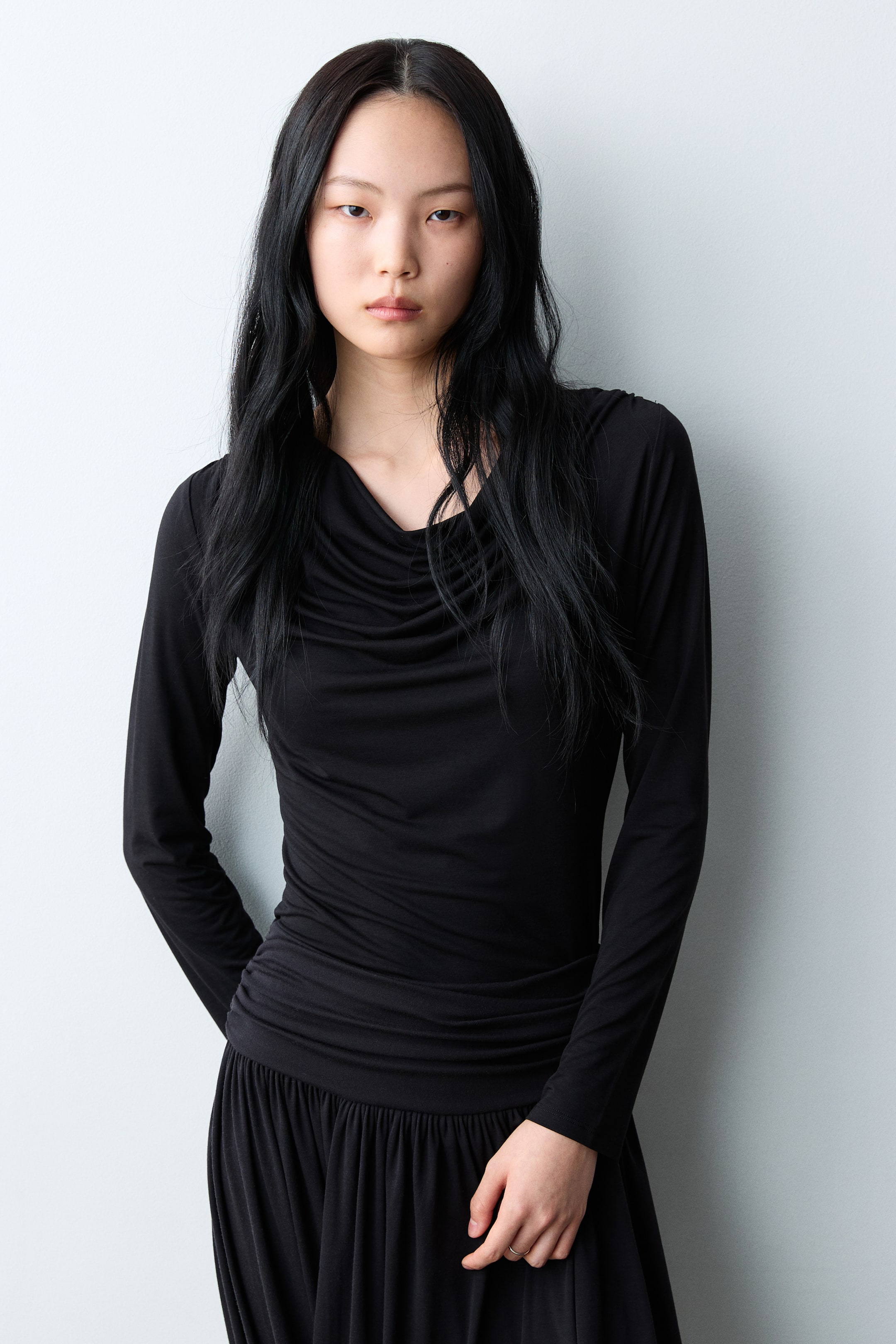 拡大画像を表示: フロントから見たブラックの長袖トップスは、ドレープのあるカウルネックラインが特徴です。ボディと袖にスリムにフィットし、ウエスト部分にはシャーリングのディテールが見られます。