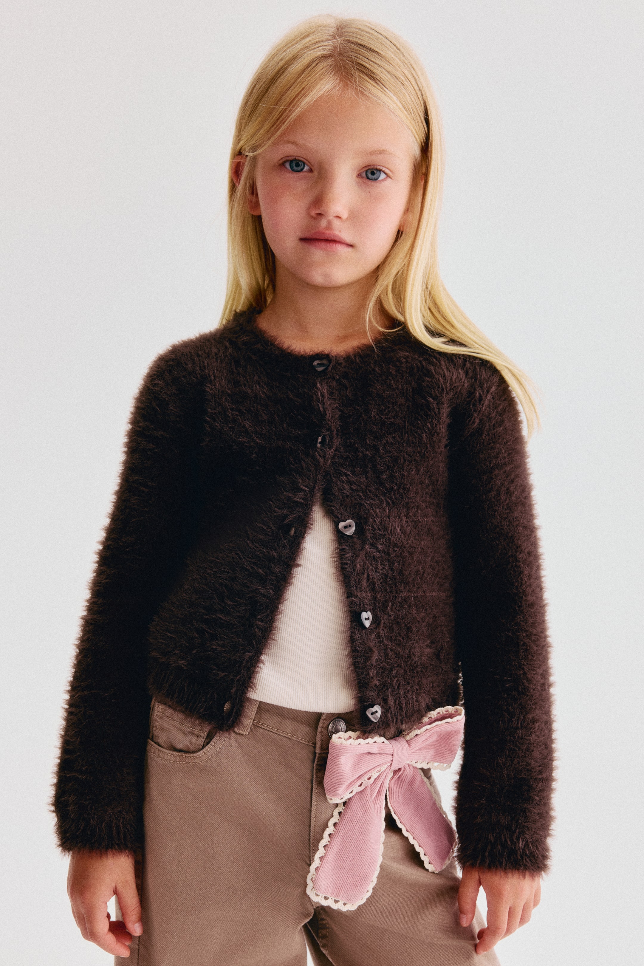 Agrandir l'image: Un cardigan marron foncé en maille douce et duveteuse est présenté de face, avec un col rond, des boutons en forme de cœur au centre et des manches longues. Le cardigan est porté par-dessus un haut blanc, mettant en valeur sa finition texturée et sa coupe décontractée.