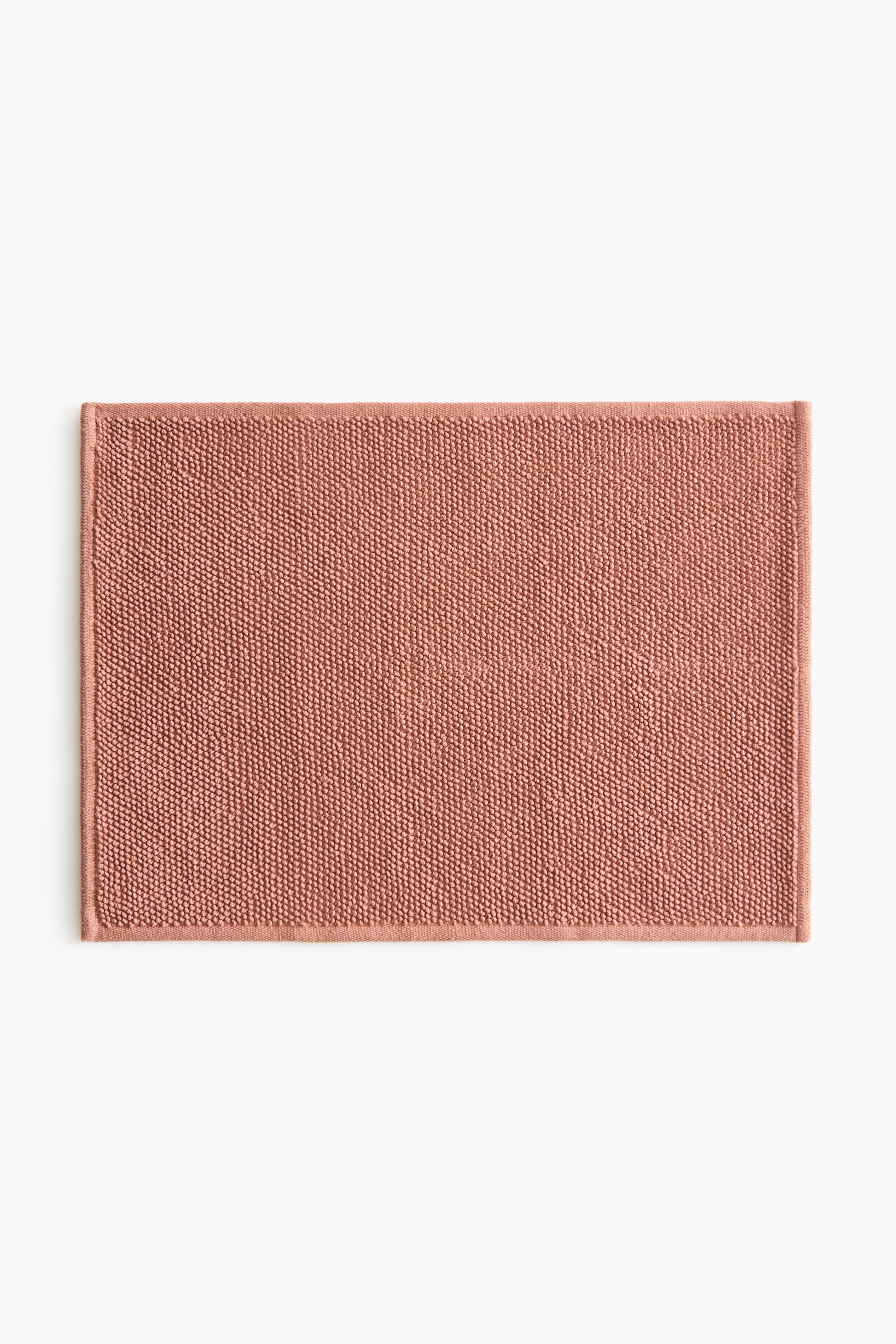 View larger image: The bath mat is rectangular and showcases its front side, highlighting a textured surface of small, dense terry loops in a soft coral hue, complete with a flat, finished edge along its perimeter.
