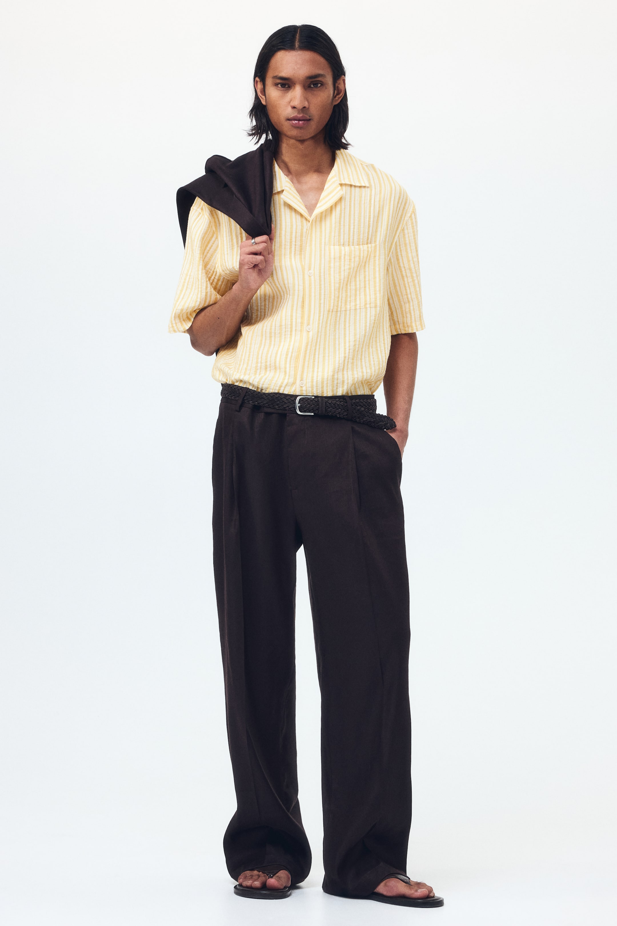 View larger image: A yellow vertically striped short-sleeve shirt with a camp collar and a single chest patch pocket is being showcased. Dark brown wide-leg trousers are being worn, featuring front pleats and an elasticated waist cinched by a plaited belt.