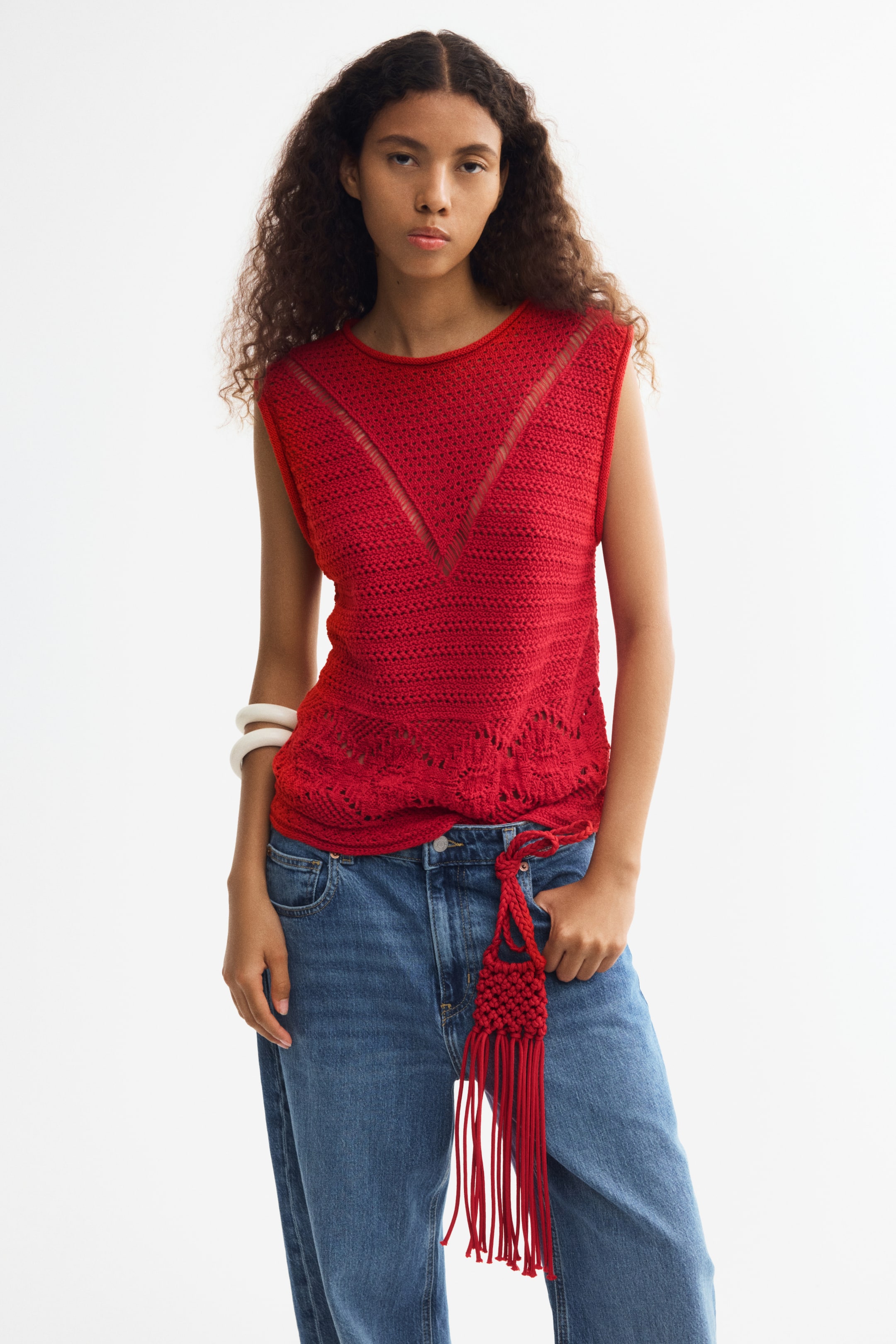 Ver imagen más grande: La modelo lleva un top rojo sin mangas de punto con aspecto de crochet, con cuello redondo, bordes vueltos y detalles calados. El top se combina con vaqueros azules y un cinturón rojo a juego con flecos.