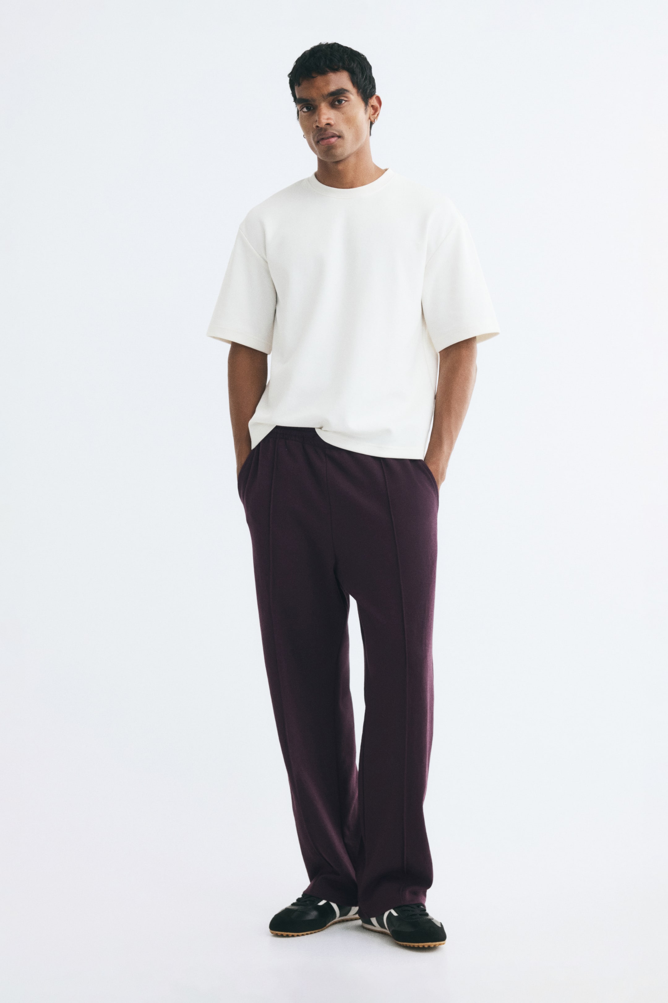 View larger image: Dark purple relaxed-fit joggers with straight legs and stitched front creases are worn by the model, who is standing with hands in the side pockets. The joggers are paired with a white short-sleeved T-shirt and black sneakers.
