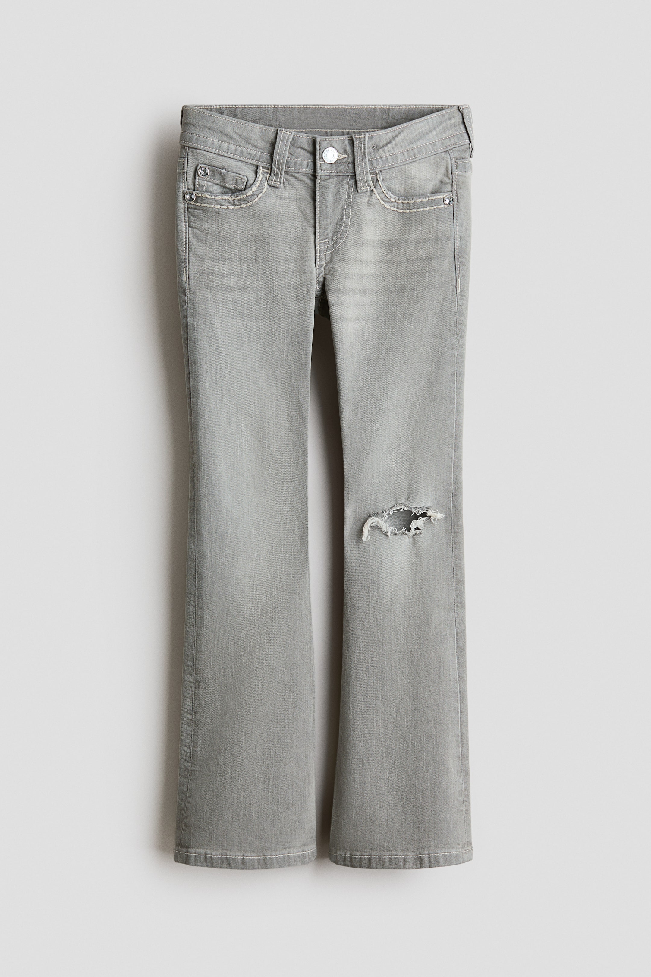 Grotere afbeelding bekijken: Grijze jeans worden van voren getoond, met een uitlopende pijp, witte contrasterende stiksels op de naden en zakken, een knoop- en ritssluiting, riemlussen en een versleten scheur op de rechterknie.