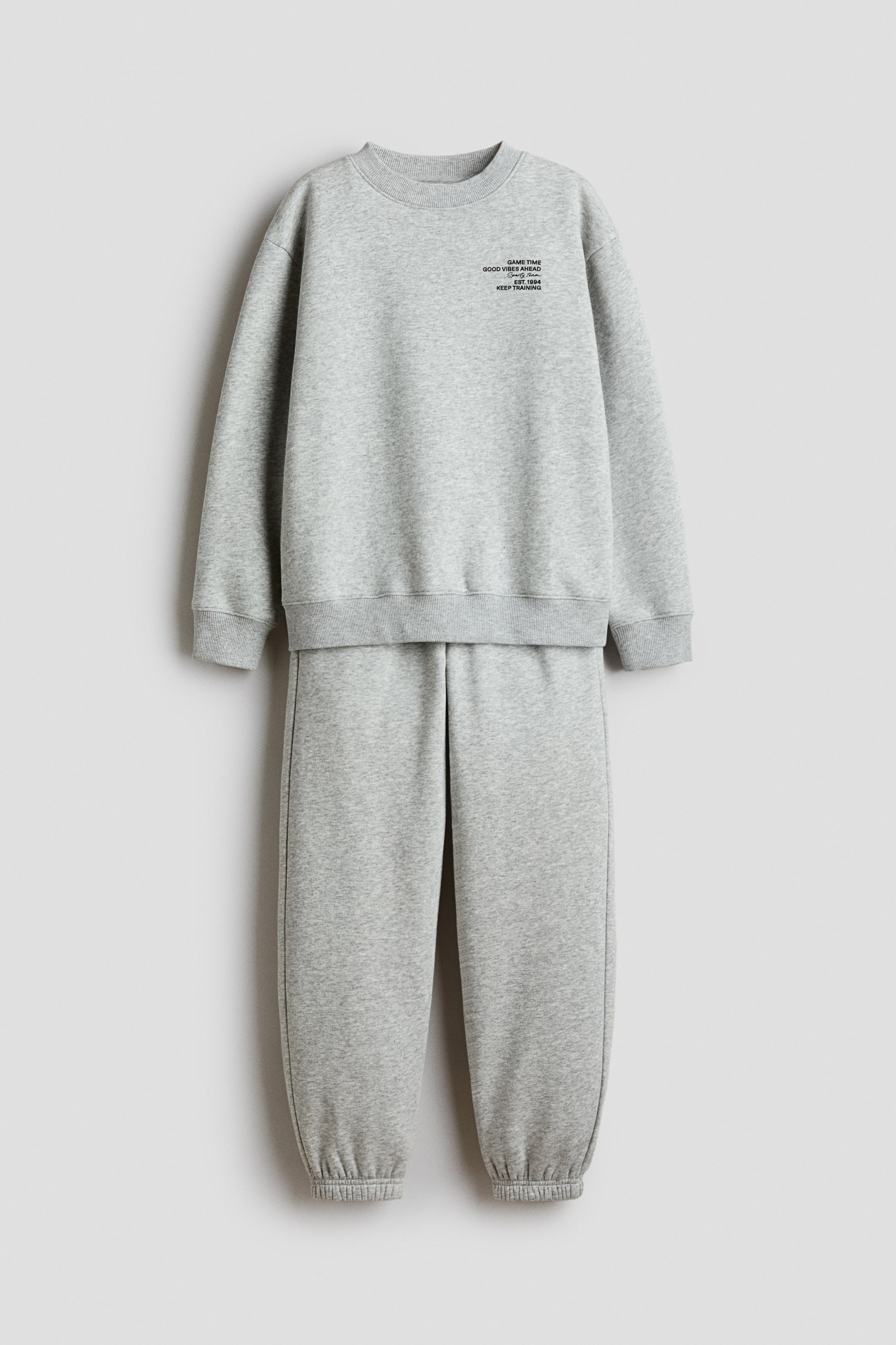 View larger image: A light grey sweatshirt and jogger set is hanging front-facing, showcasing a coordinated lounge design. The long-sleeved sweatshirt features a crew neck, ribbed trims, and small black text on the left chest, while the matching joggers feature gathered elastic cuffs at the ankles.