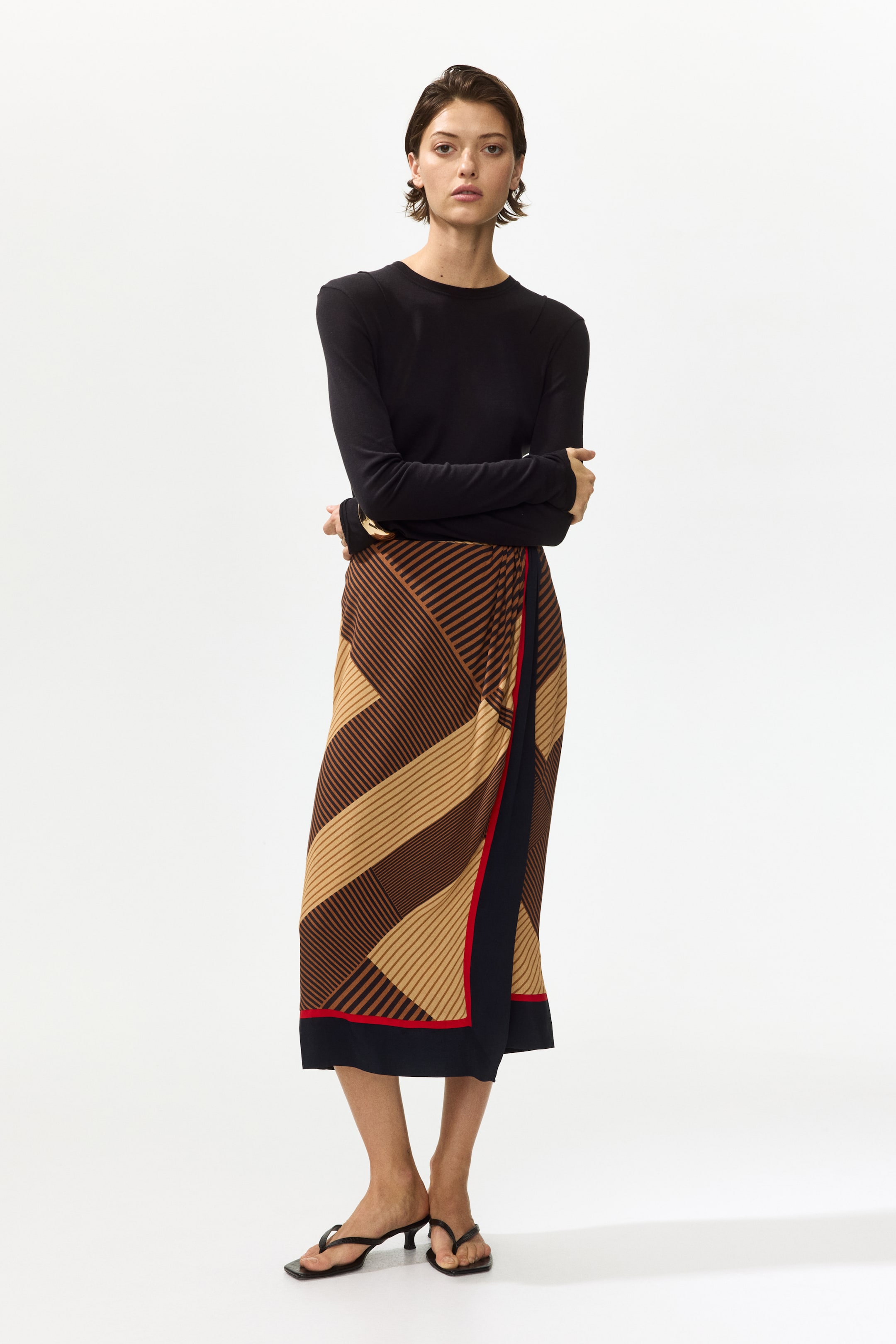 Büyük resmi görüntüle: Midi sarmal etek, ten ve kahverengi tonlarında kesişen çapraz çizgilerden oluşan geometrik bir desen sergiliyor. Üst üste binen ön paneli ve etek ucunda siyah ve kırmızı bir sınırla dikkat çekiyor.