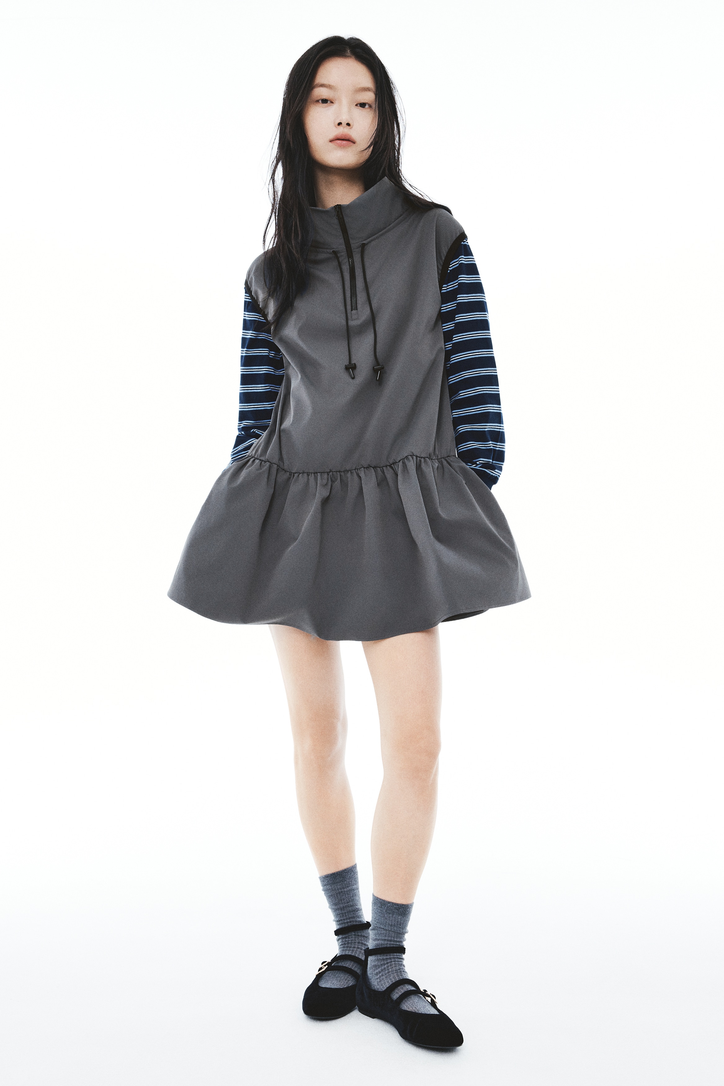 A sleeveless mini dress in dark grey twill is shown facing front, featuring a high stand collar with a quarter-zip closure and adjustable drawstrings. The dress features black elasticated trim along the armholes and a gathered, dropped waist that flares out towards the hem.