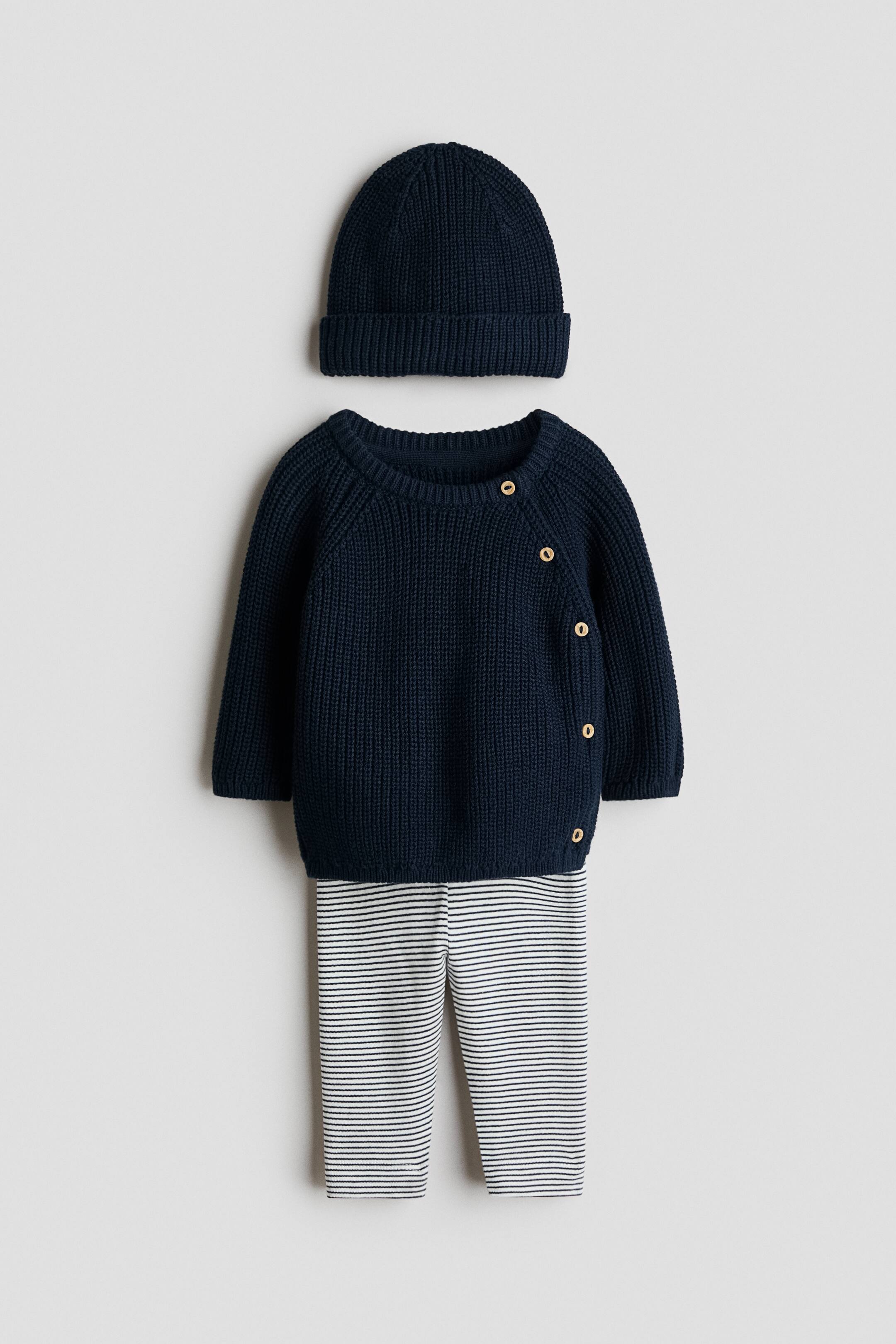 View larger image: A three-piece baby outfit featuring a dark blue ribbed knit beanie and a matching dark blue ribbed knit sweater with raglan sleeves. The sweater features an asymmetrical front closure with light brown circular buttons on the left shoulder and side, and is layered over white pants with horizontal dark blue stripes.