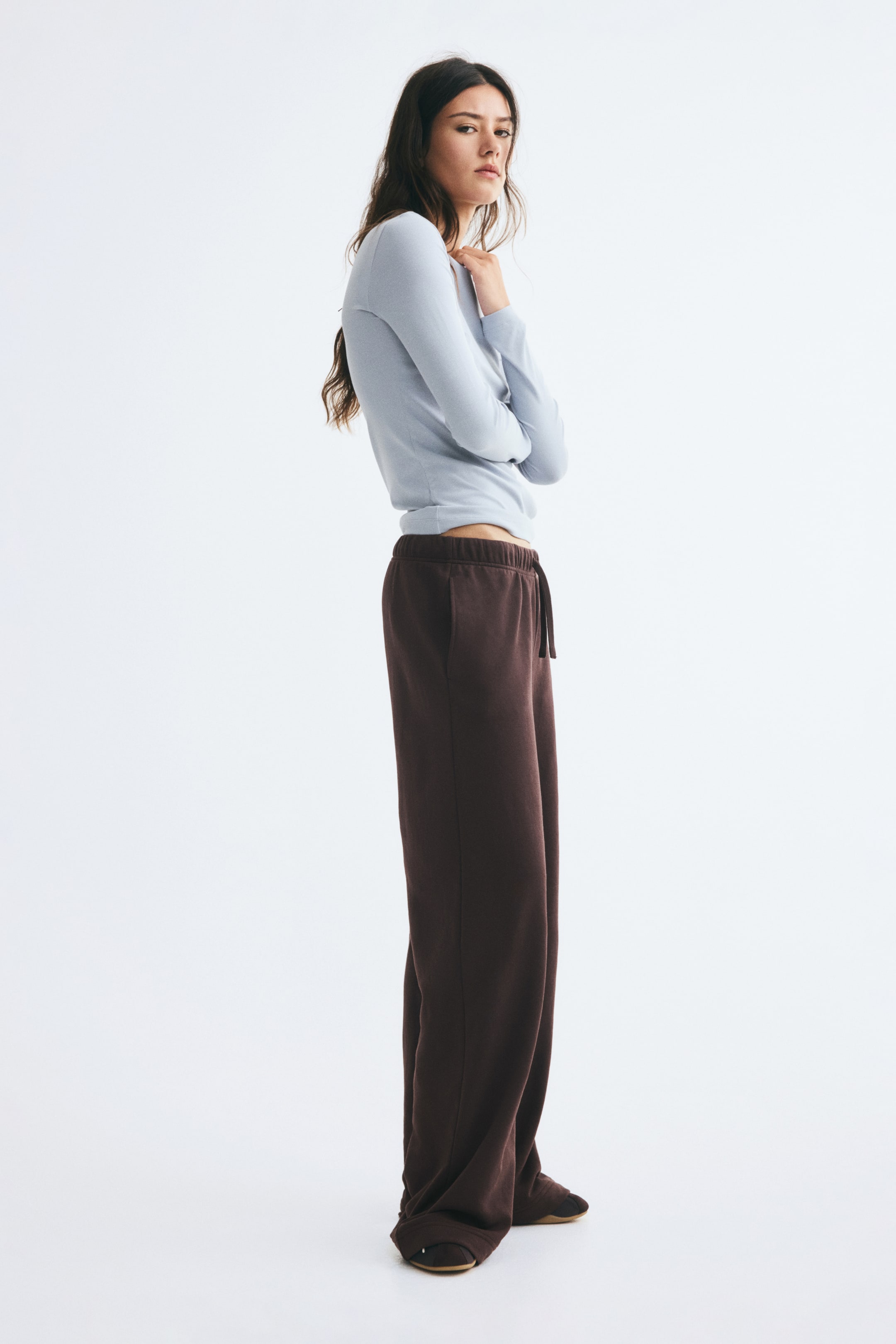 View larger image: Track pants in dark brown sweatshirt fabric, featuring an elasticized waistband with a drawstring and a straight-leg silhouette, are worn by the model standing in profile. The pants are styled with a light blue long-sleeve top.