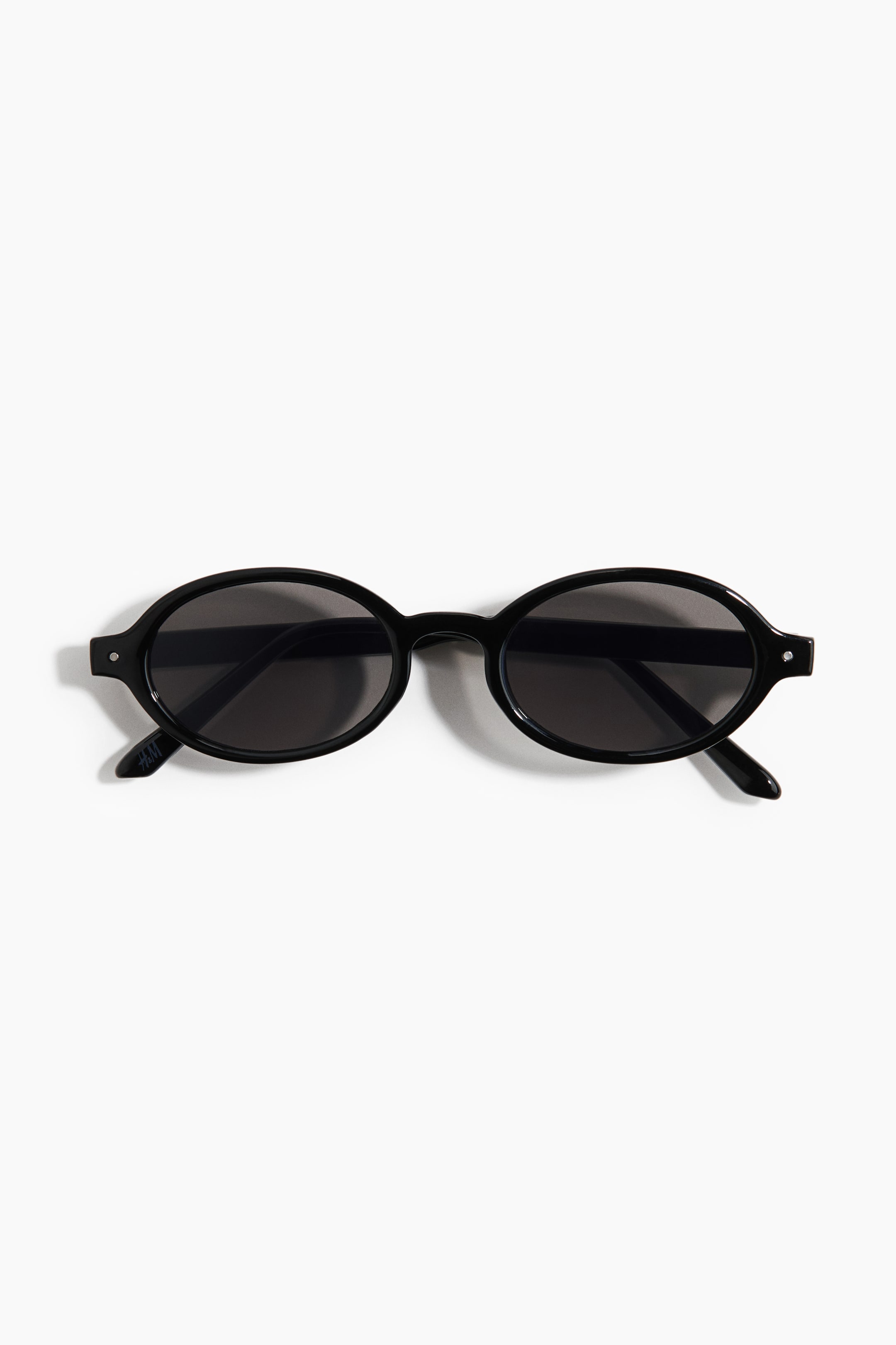 Agrandir l'image: Lunettes de soleil ovales noires orientées de FACE, avec des montures épaisses, des verres foncés et de petits clous argentés visibles sur les coins extérieurs. Les branches sont repliées vers l'intérieur, avec un petit logo "H&M" apparaissant sur la branche gauche.