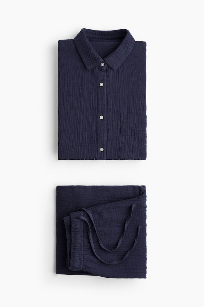 A pair of navy pyjamas, made from soft cotton muslin, is presenting a folded shirt and folded bottoms. The shirt is featuring a collar, light-coloured buttons down the front, and a patch chest pocket, while the bottoms are displaying an elasticated, drawstring waist with straight legs. - 1